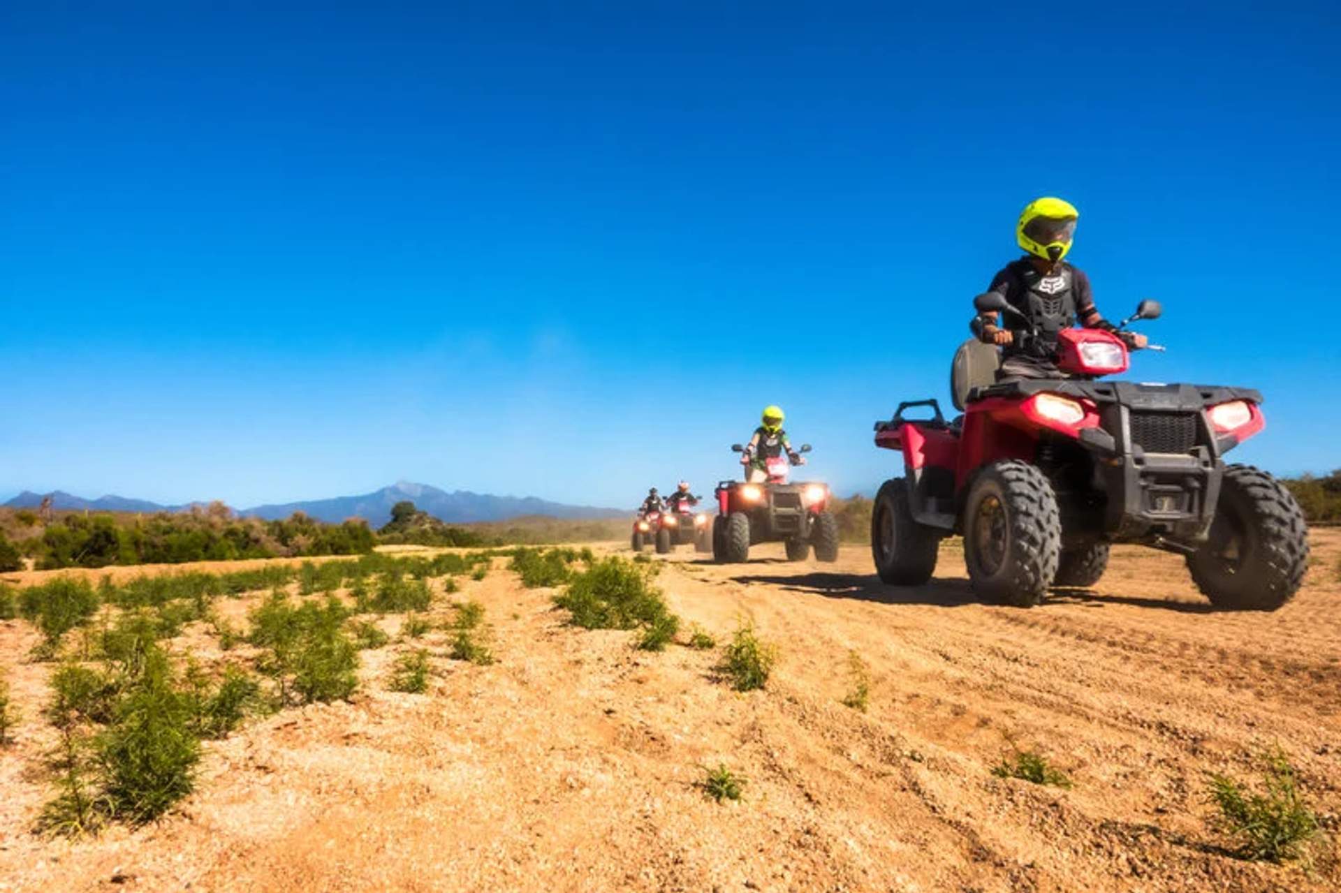 Grupo de personas montando cuatrimotos rojas por un sendero desértico bajo un cielo azul despejado, con montañas al fondo.