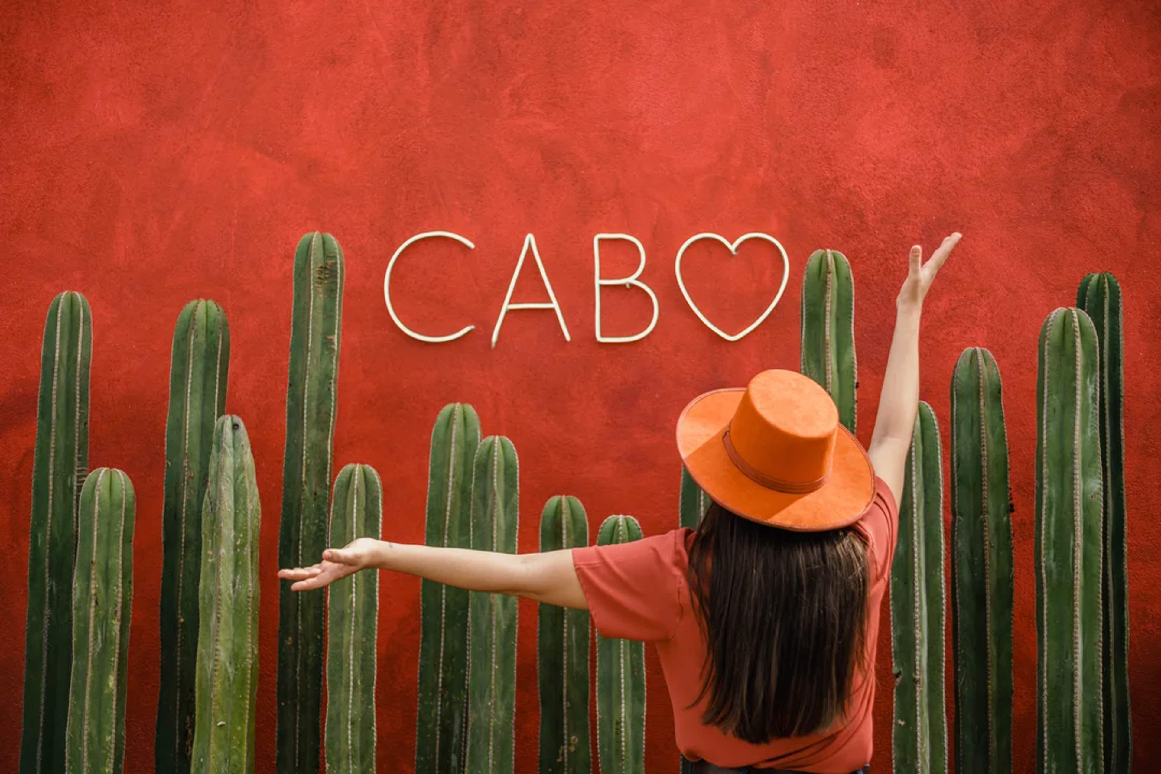 Viajera posa frente a un muro rojo con cactus y un letrero luminoso que dice “Cabo”.