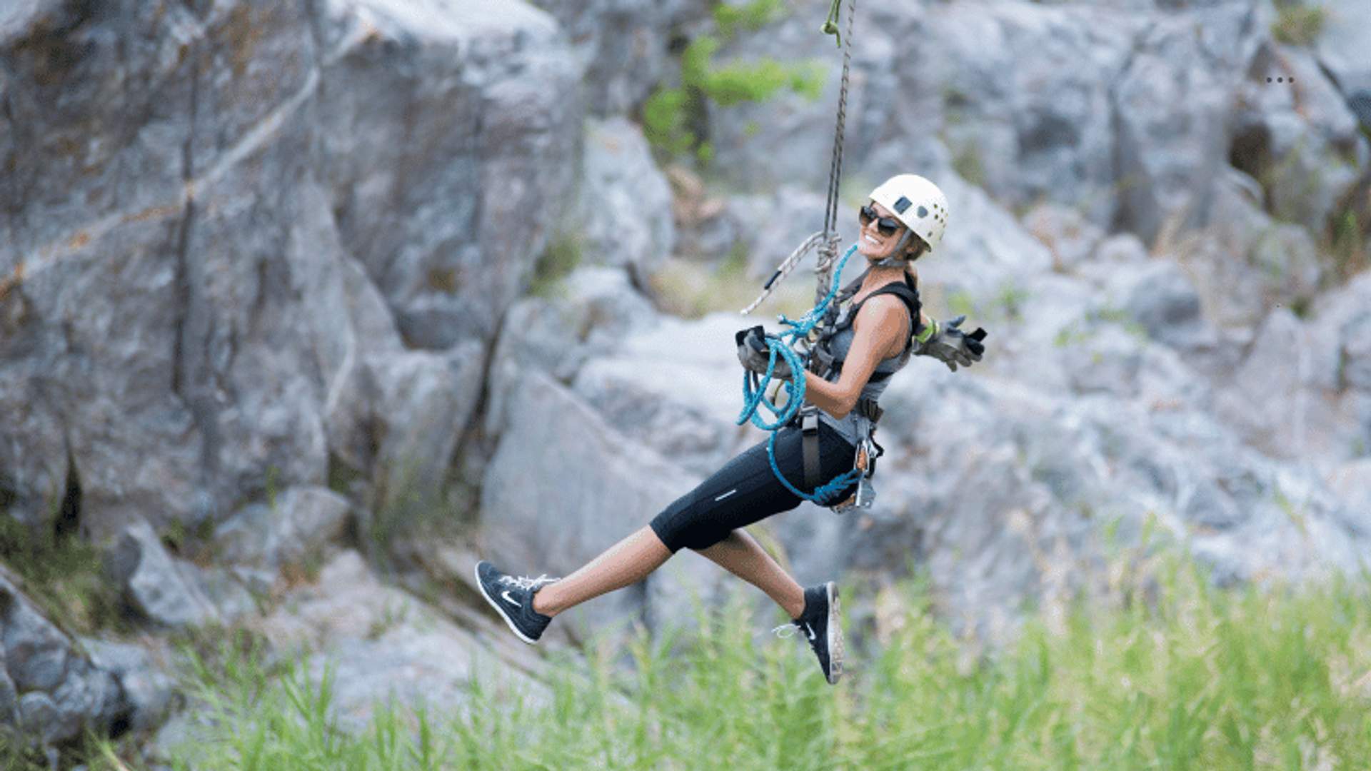 Una mujer disfruta de una tirolesa por un cañón rocoso en Cabo, sonriendo y suspendida en el aire, mostrando una cita romántica y aventurera.