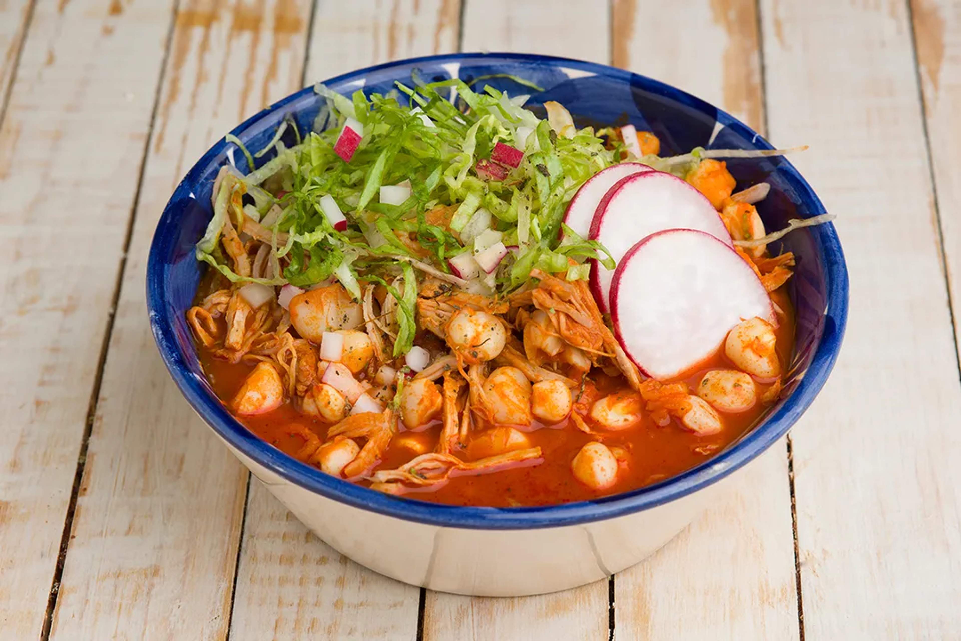 Traditional red pozole with chicken, hominy, lettuce, radish, and onion in a blue bowl.