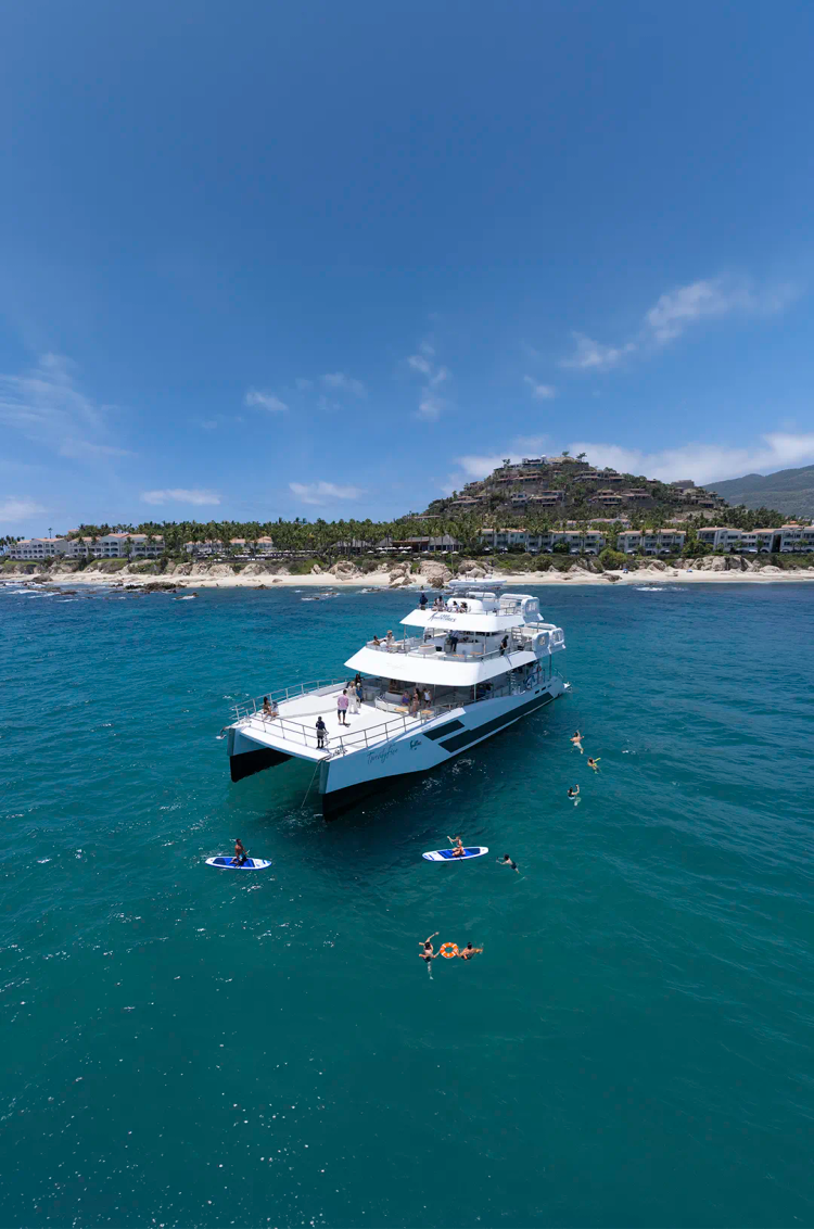 Luxury catamaran in Los Cabos with guests snorkeling and paddle boarding in turquoise waters.