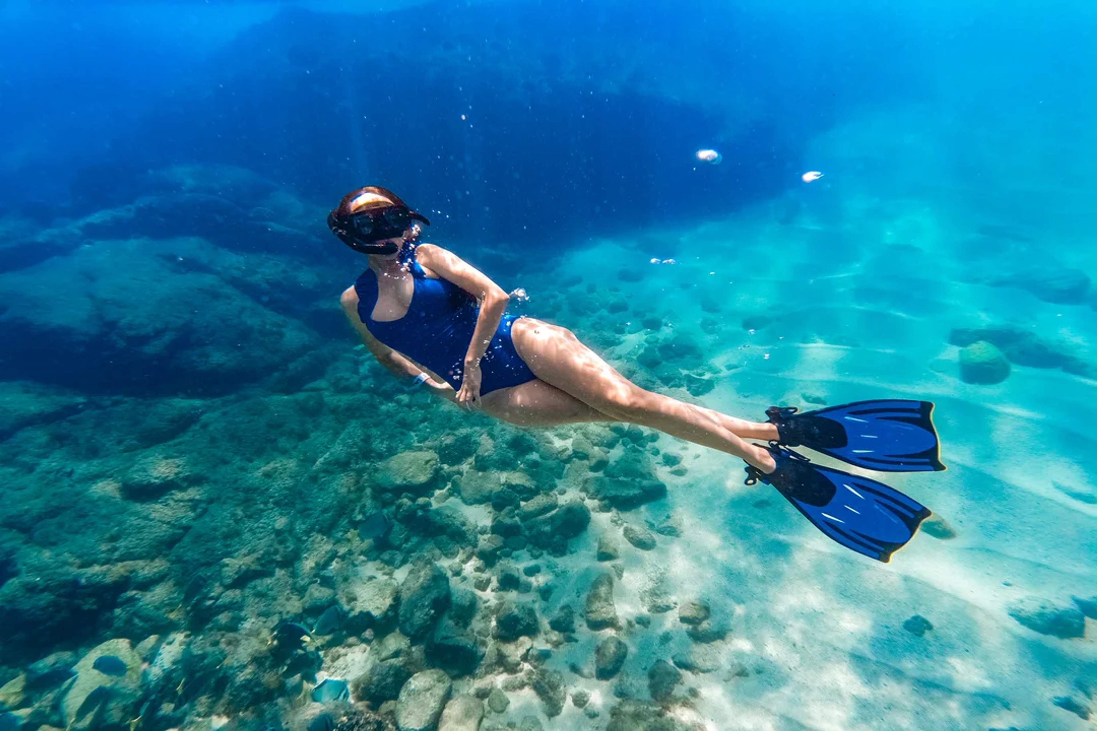 A woman in a blue swimsuit snorkels in crystal-clear waters, exploring an underwater rocky landscape.