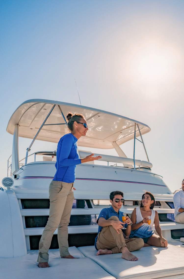 Una guía habla con un grupo de personas disfrutando de bebidas en un yate cerca de acantilados rocosos en Cabo San Lucas.