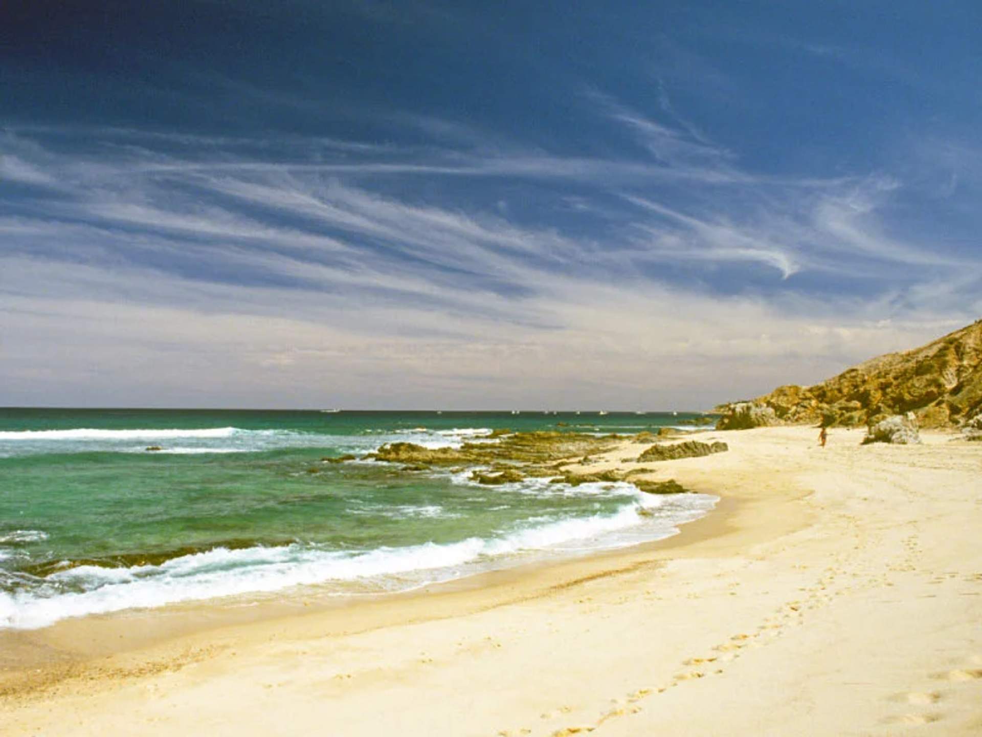 Playa tranquila con arena dorada, rocas y cielo con nubes onduladas en un día soleado.