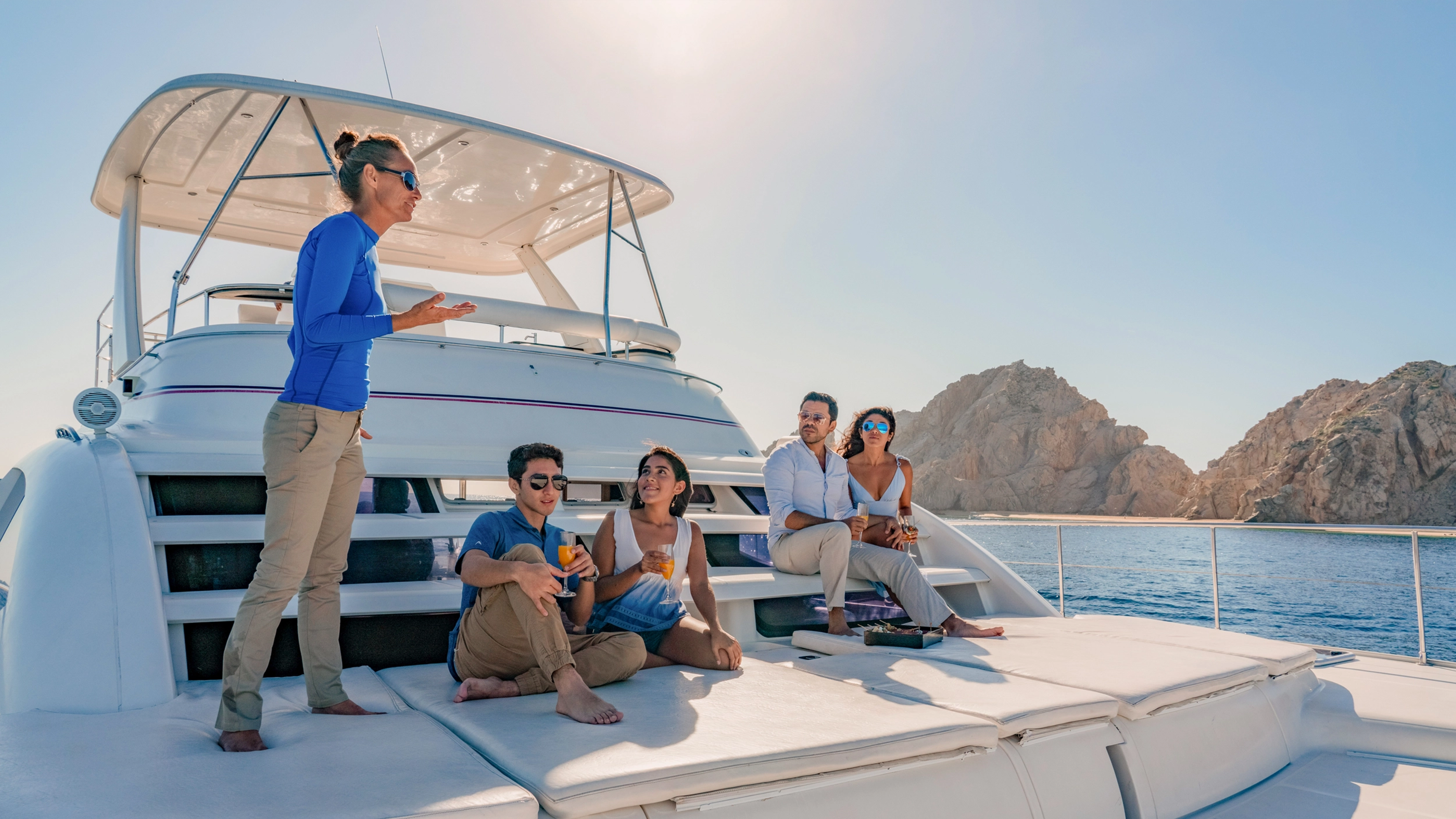 Grupo de amigos se relajan en un yate de lujo, disfrutando bebidas bajo el sol con acantilados de fondo.