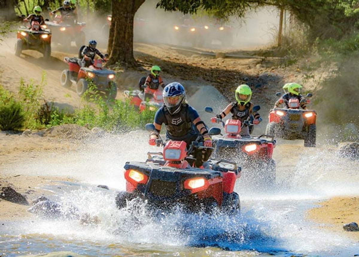 A group of riders on red ATVs splashing through water on a dirt trail, wearing helmets and protective gear in Cabo.