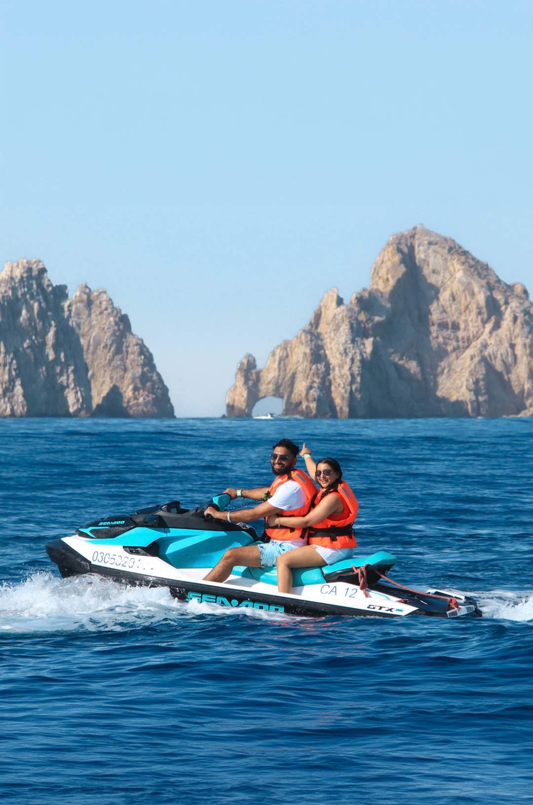 A couple rides a jet ski in the ocean with large rock formations in the background, both wearing orange life jackets.