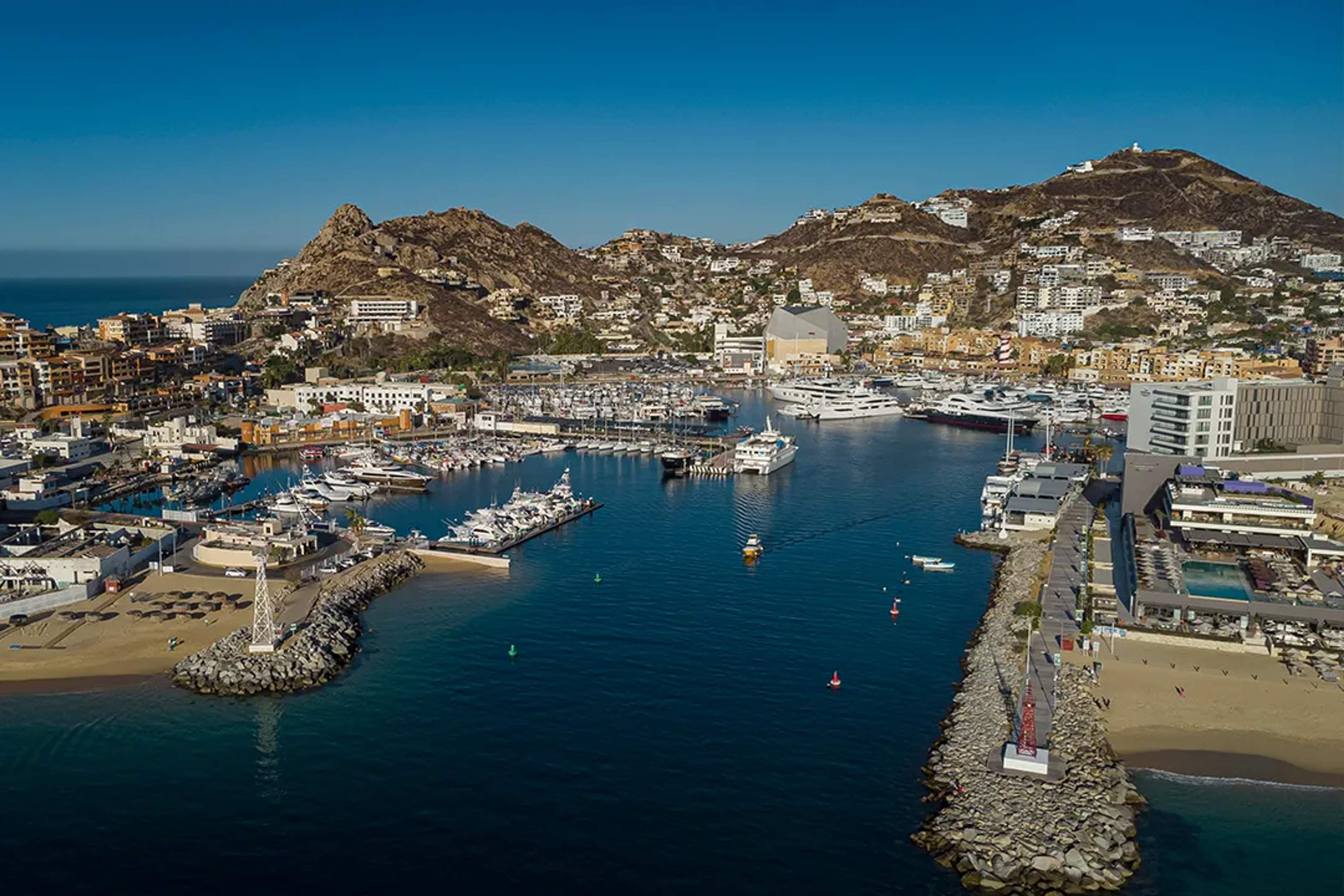 Vista aérea de la marina de Cabo San Lucas, con yates, hoteles y colinas bajo un cielo despejado.