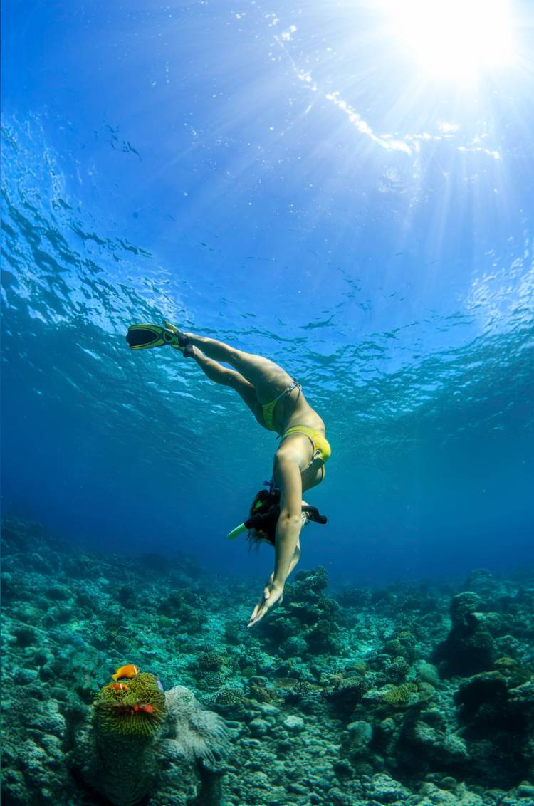 Woman in a yellow bikini diving underwater, surrounded by vibrant coral and marine life, with sunlight streaming down.