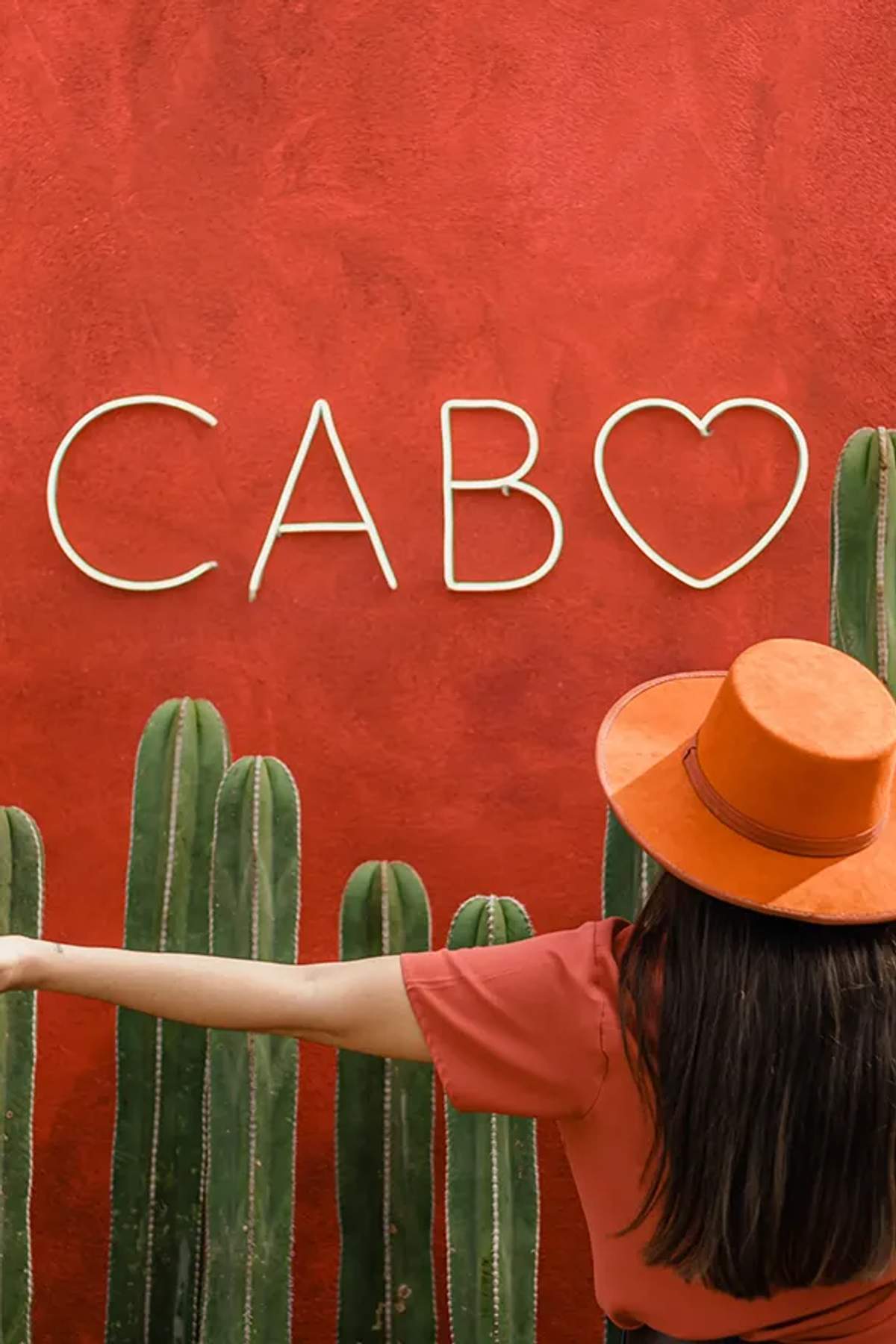 Woman in orange hat at cactus mural with CABO sign, iconic photo spot in Los Cabos, Mexico.