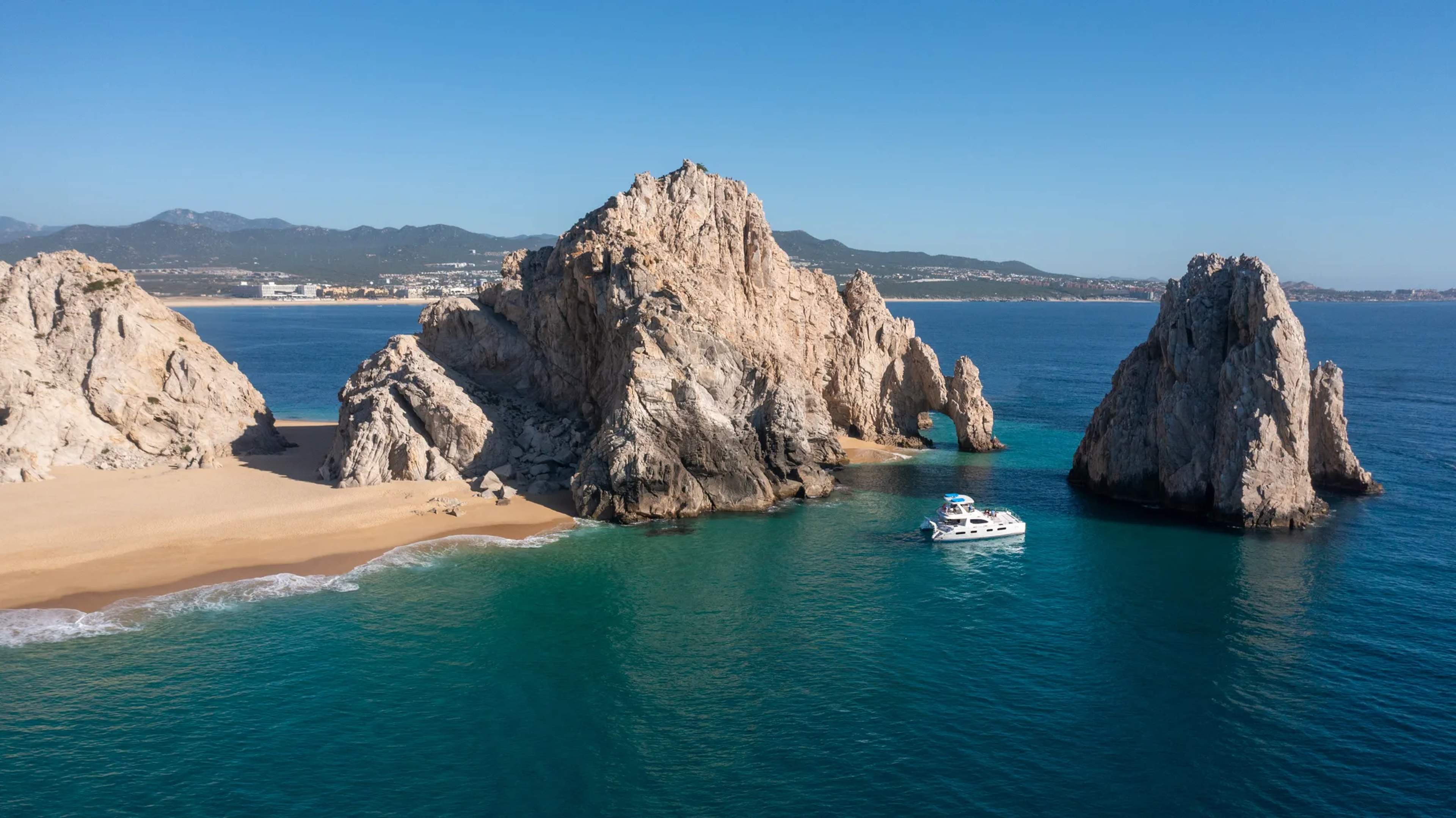 A white boat anchors near Cabo San Lucas rock formations, with turquoise waters and golden sand beach in the foreground.