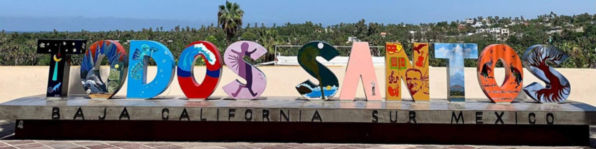 Colorful "Todos Santos" sign in Baja California Sur, Mexico, with vibrant letters and scenic palm trees and hills in the background.