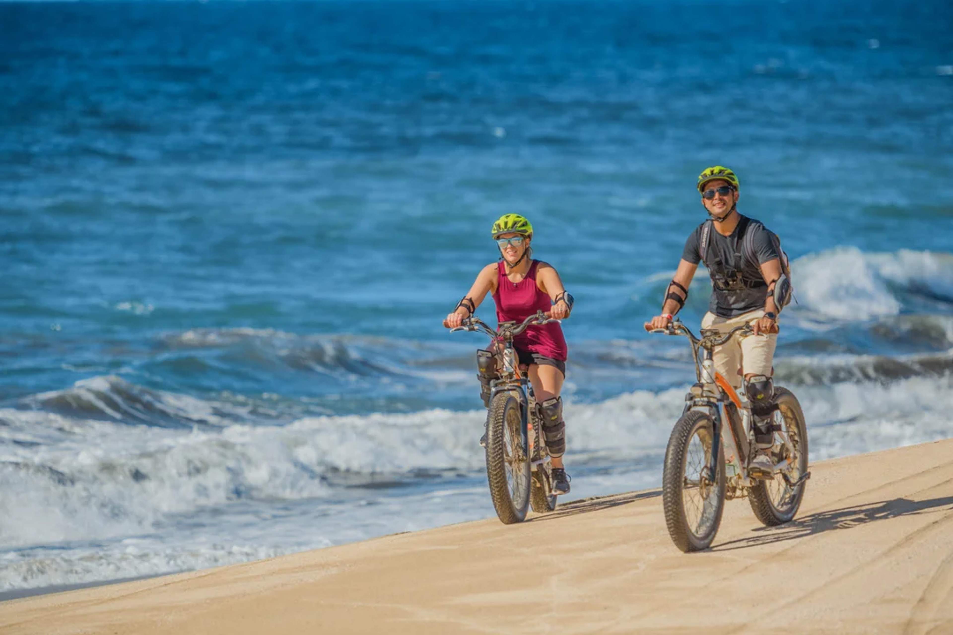 Couple rides electric bikes along the sandy beach, smiling under the sun by the ocean waves.