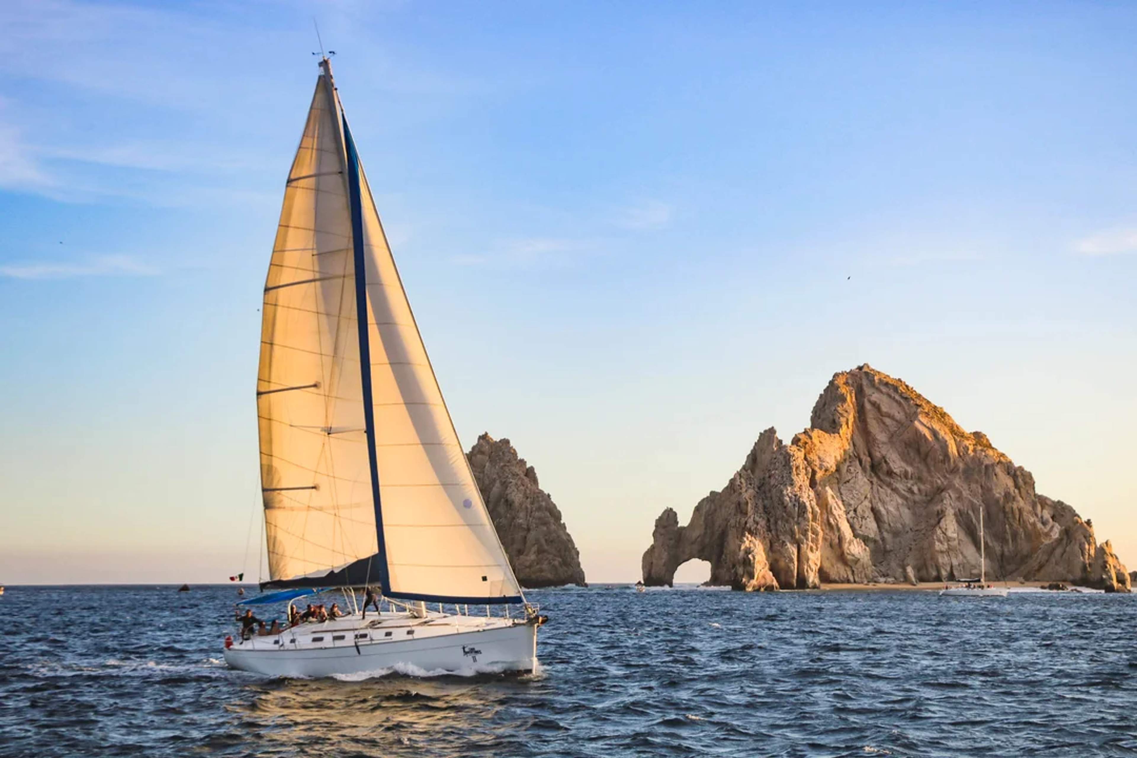 Paseo en velero al atardecer frente al icónico Arco de Cabo, con luz dorada sobre el mar y rocas.