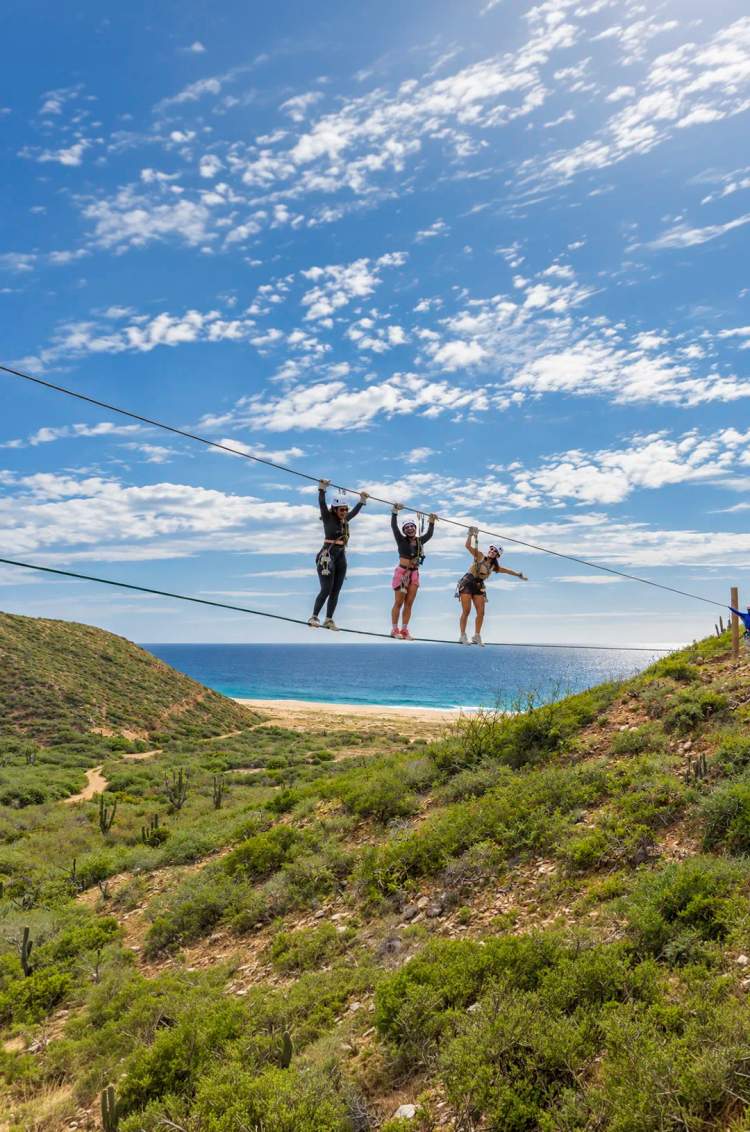 Three women cross a suspended rope bridge at Tierra Sagrada with views of the Pacific Ocean in Los Cabos.