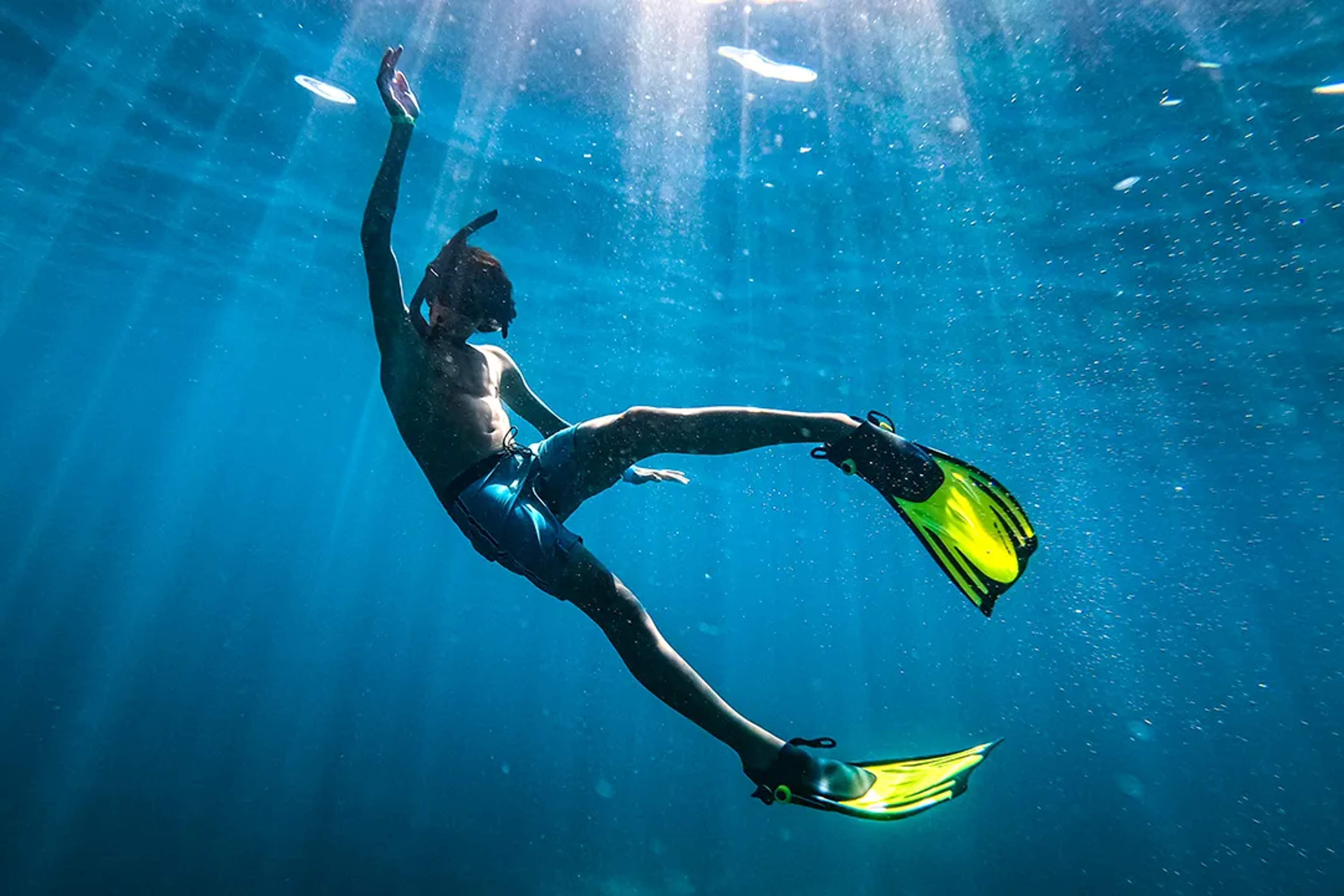 Persona flota bajo el agua con aletas amarillas, rodeada de rayos de sol y burbujas marinas.