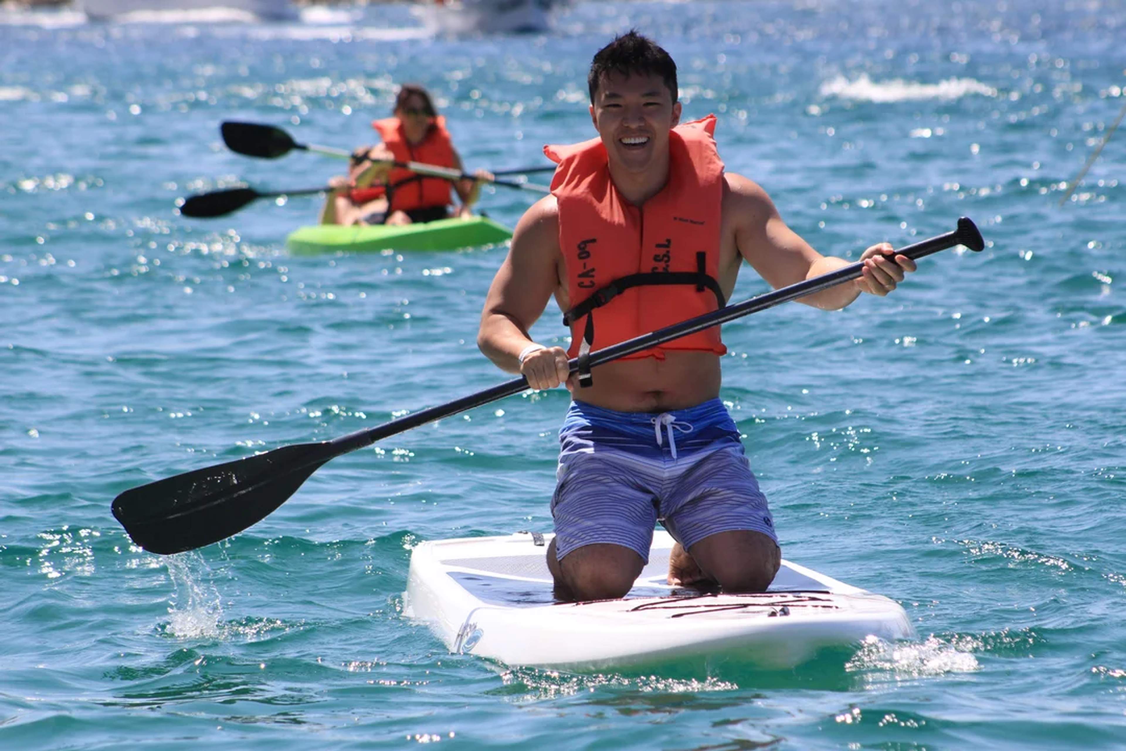 Un hombre disfruta del paddleboard en aguas cristalinas, sonriendo bajo el sol, mientras un kayakista lo sigue al fondo.