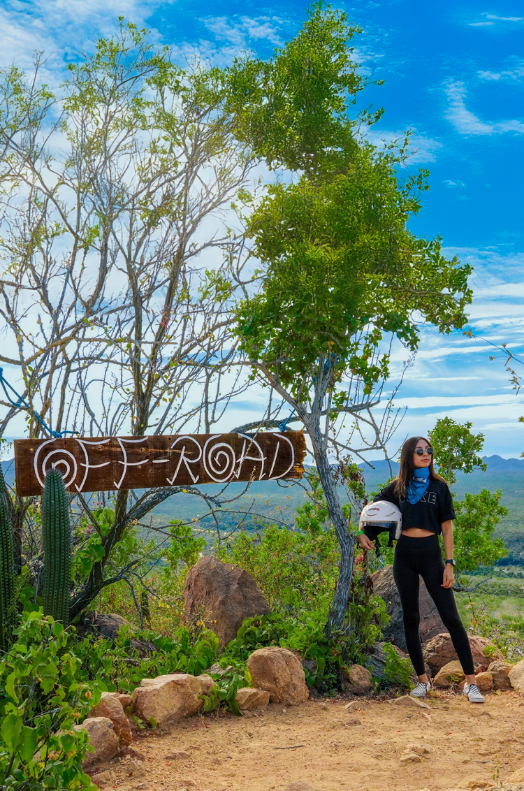 Mujer de negro junto a un letrero de "Off-Road", sosteniendo un casco, rodeada de plantas desérticas y un paisaje montañoso.