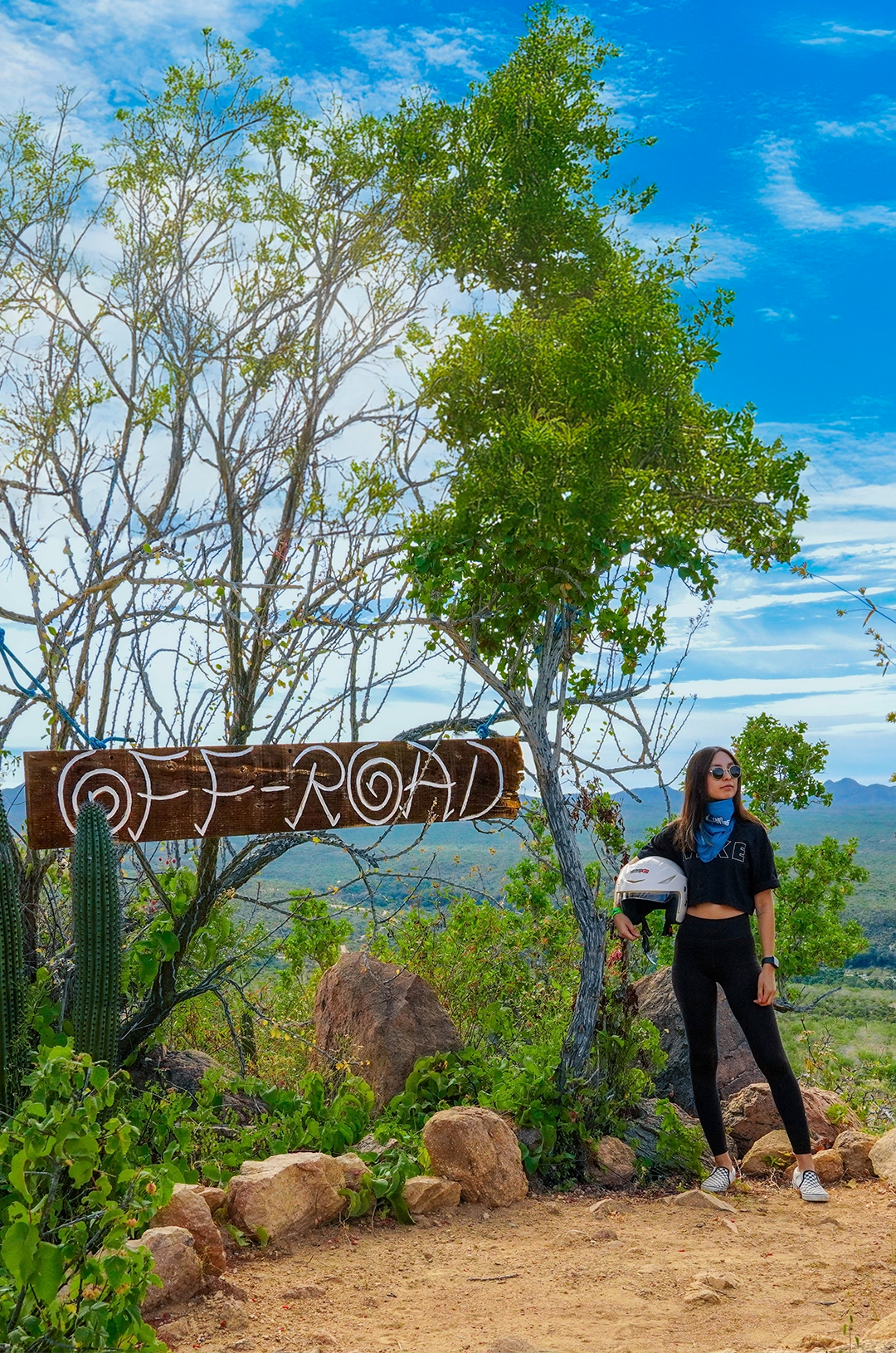 Woman stands beside an "Off-Road" sign, holding a helmet, surrounded by desert plants and a scenic mountainous backdrop.