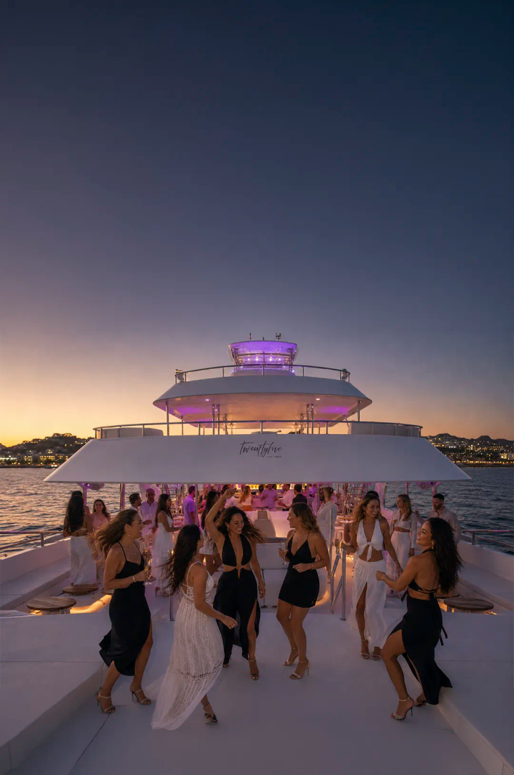 Invitados bailando en un catamarán de lujo al atardecer en Cabo, con vista al mar y luces de la ciudad.