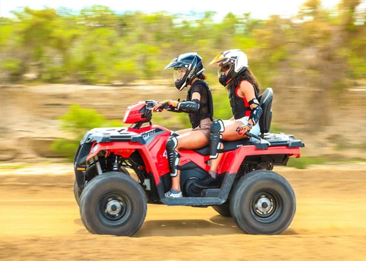 Two riders on a red ATV, wearing helmets and protective gear, driving on a dirt trail in Cabo.