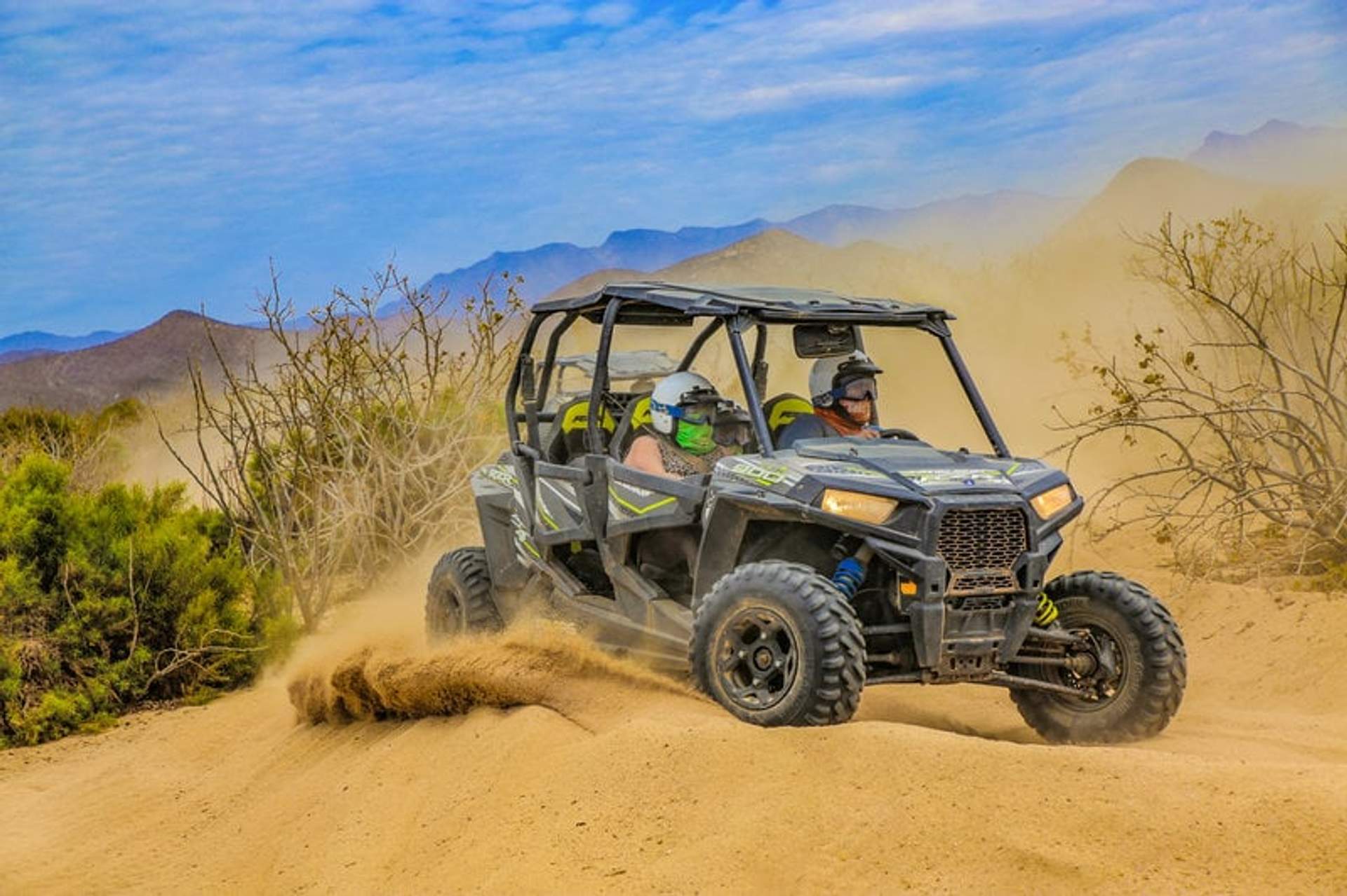 Dos personas conducen un vehículo todoterreno por un sendero arenoso en el desierto, levantando una nube de polvo mientras avanzan.