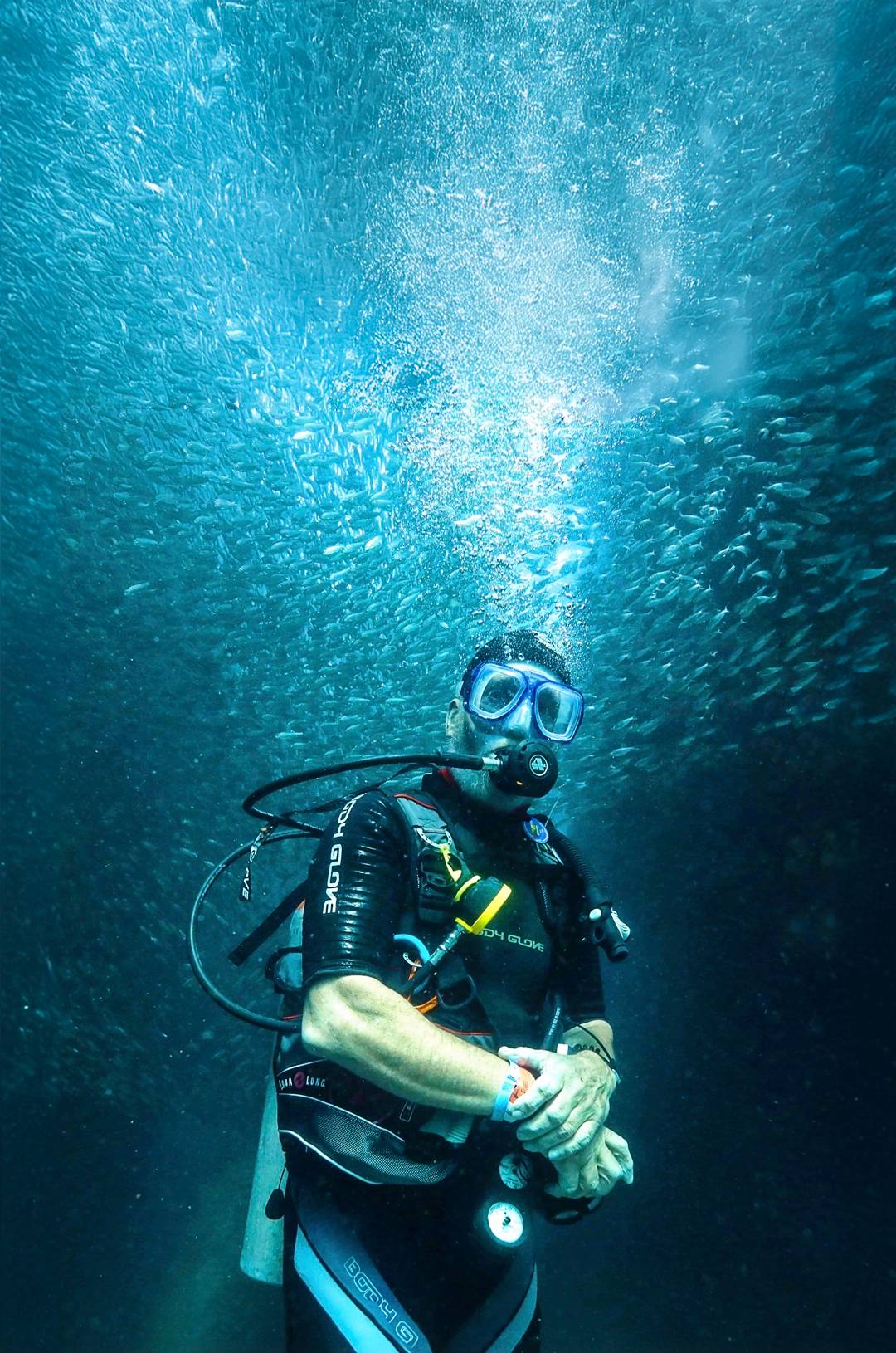 Buzo rodeado de un brillante banco de peces, con burbujas ascendiendo en el agua azul profunda.