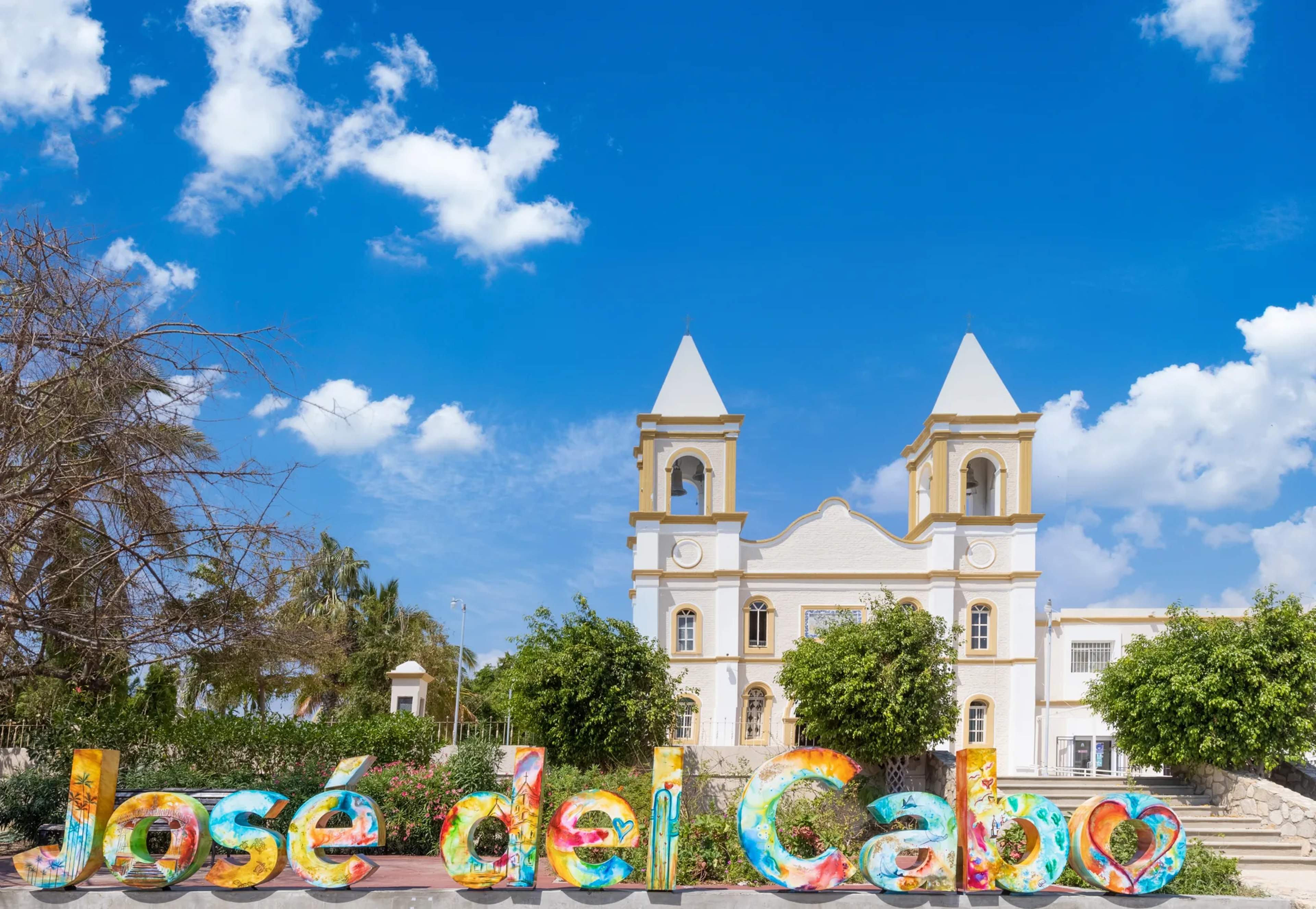 Colorful ground lettering spelling San Jose del Cabo in front of a white colonial church with twin bell towers