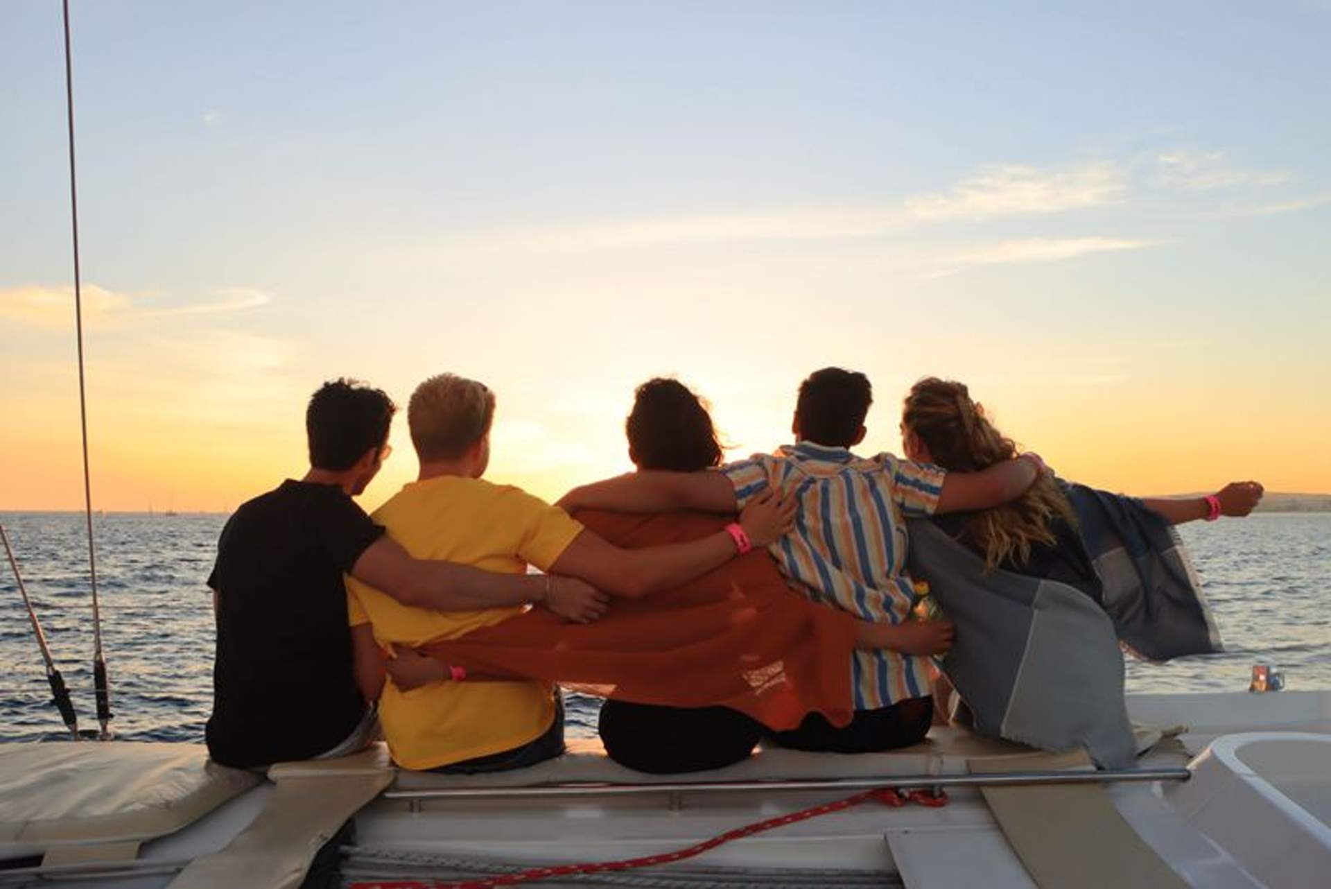 Cinco amigos sentados en un barco, abrazados, viendo la puesta de sol sobre el océano.