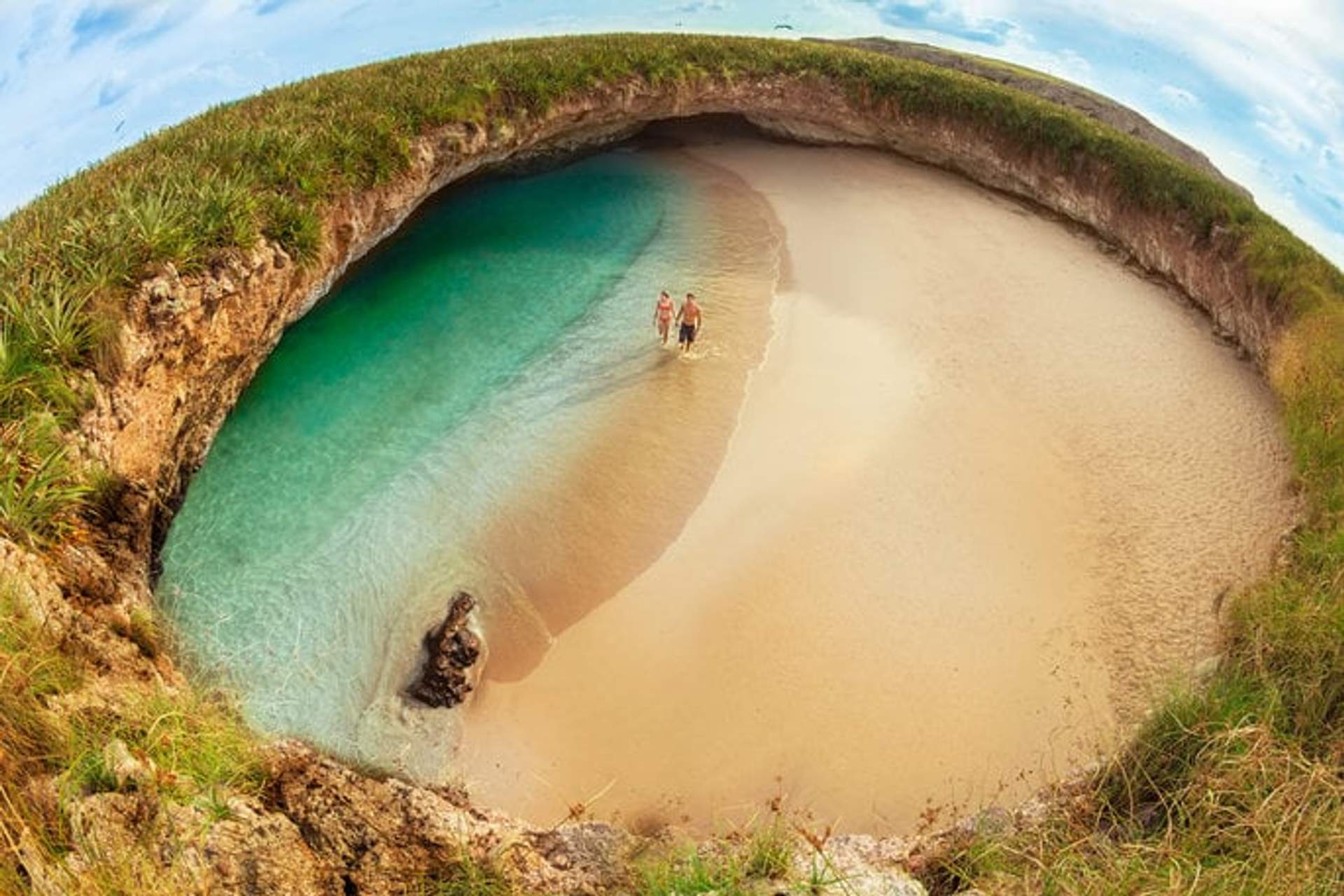 Dos personas caminan por una playa escondida con arena dorada y agua turquesa, rodeada por un anillo de roca y vegetación. La imagen muestra la famosa Playa del Amor en las Islas Marietas, conocida por su formación geológica única.