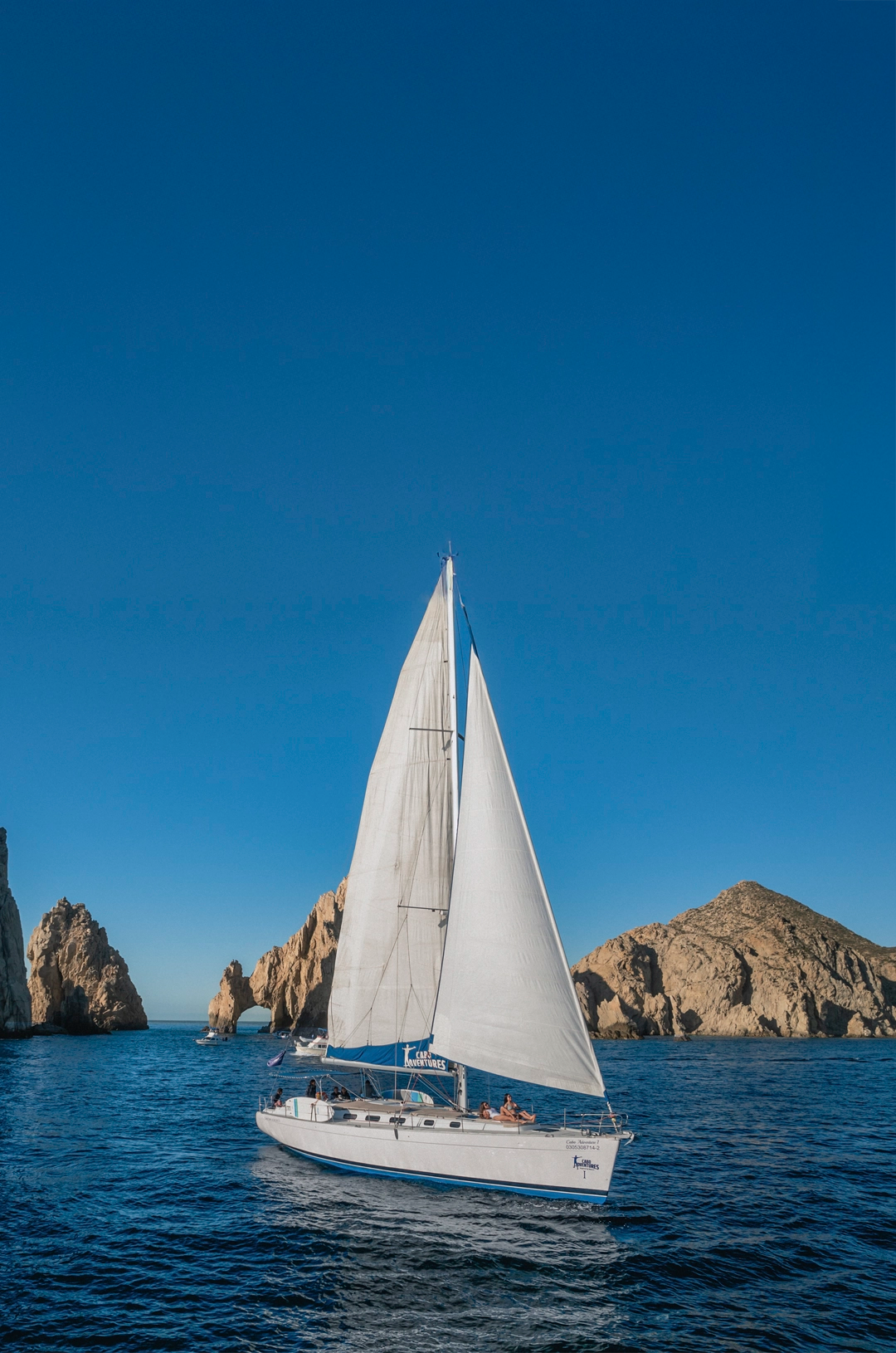 Sailboat glides near rock formations under a clear blue sky in Cabo San Lucas, with the famous arch in the background.