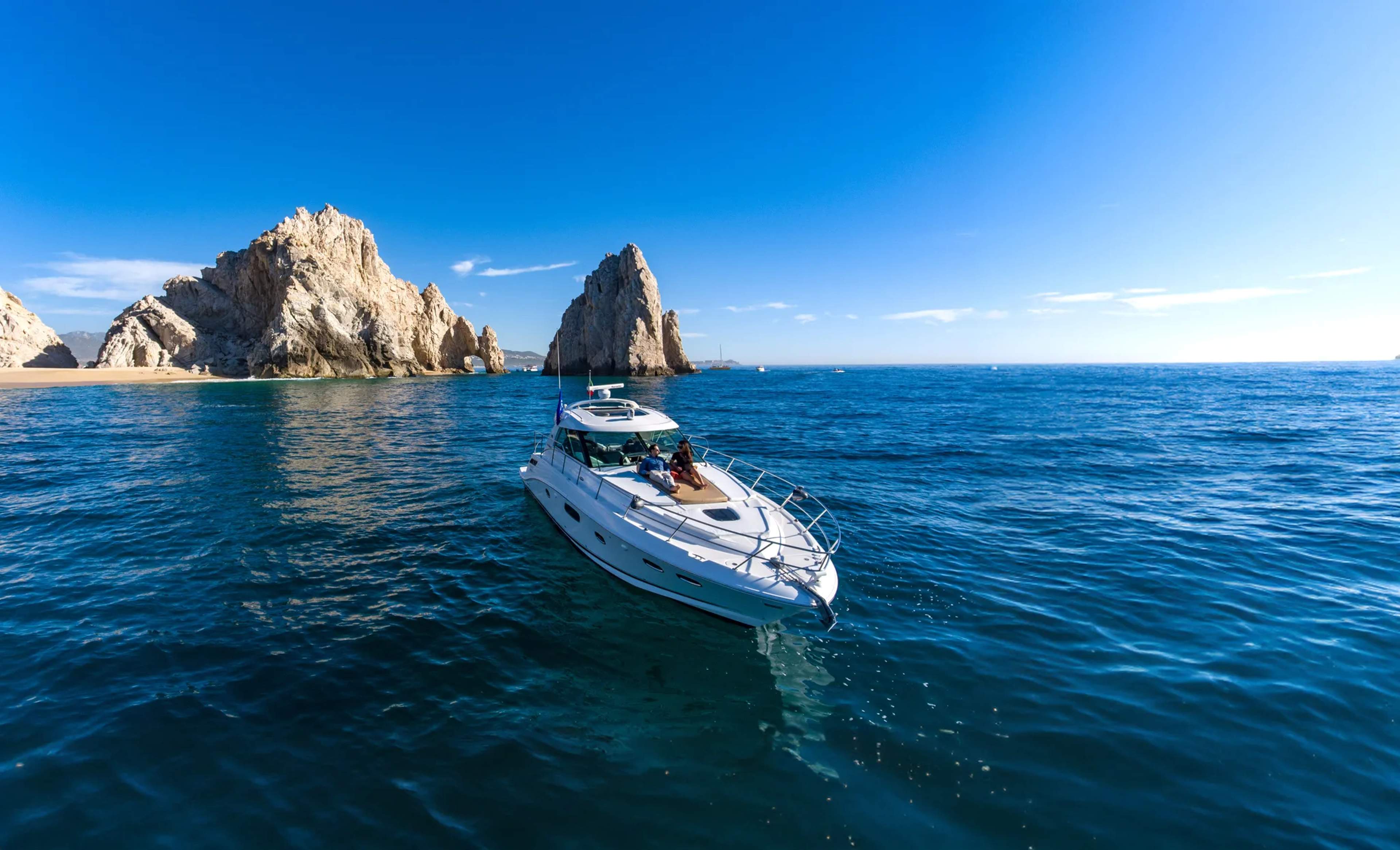 A luxury yacht anchors in clear turquoise waters beside the dramatic rocky peaks of Cabo San Lucas on a calm day.