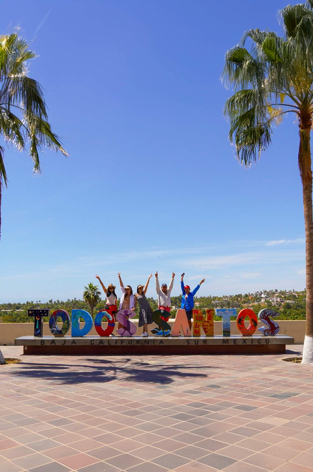Grupo saltando frente al colorido letrero de "Todos Santos" en Baja California Sur, México, con palmeras y cielo azul.