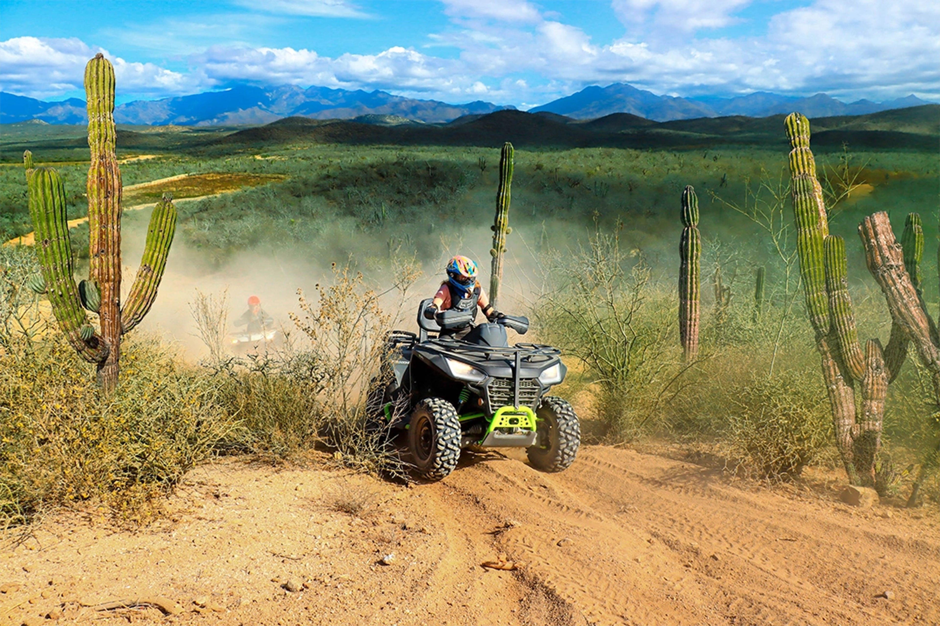 Un conductor de ATV recorre un camino polvoriento en el desierto rodeado de cactus y montañas bajo un cielo azul brillante.