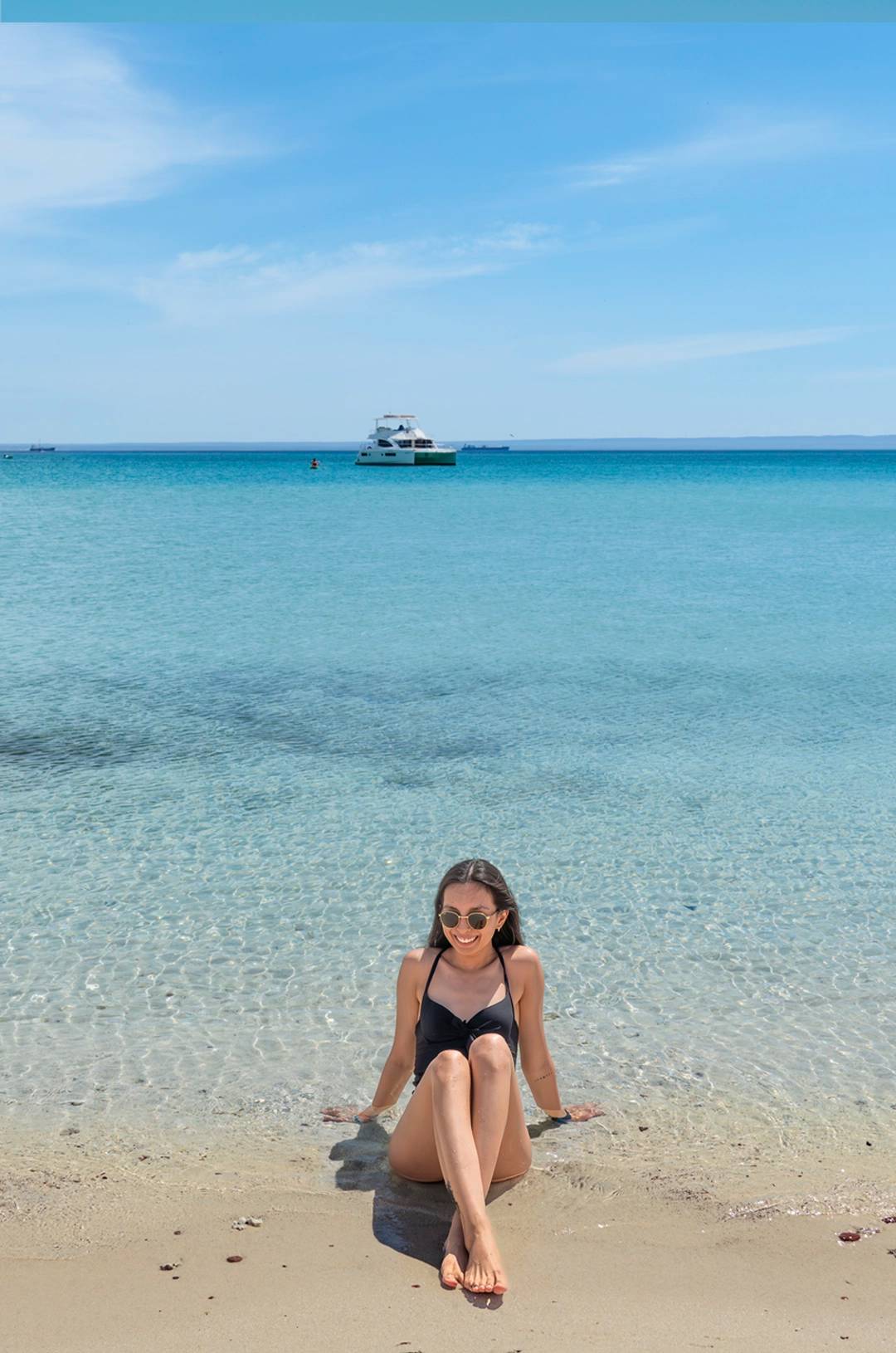 A woman in a black swimsuit sitting at the edge of the clear water on a beach, with a yacht in the distance.