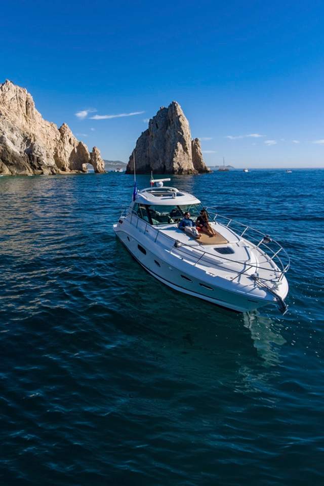 A luxury yacht cruises near the iconic rock formations of Los Cabos under a clear blue sky, with two people relaxing on deck