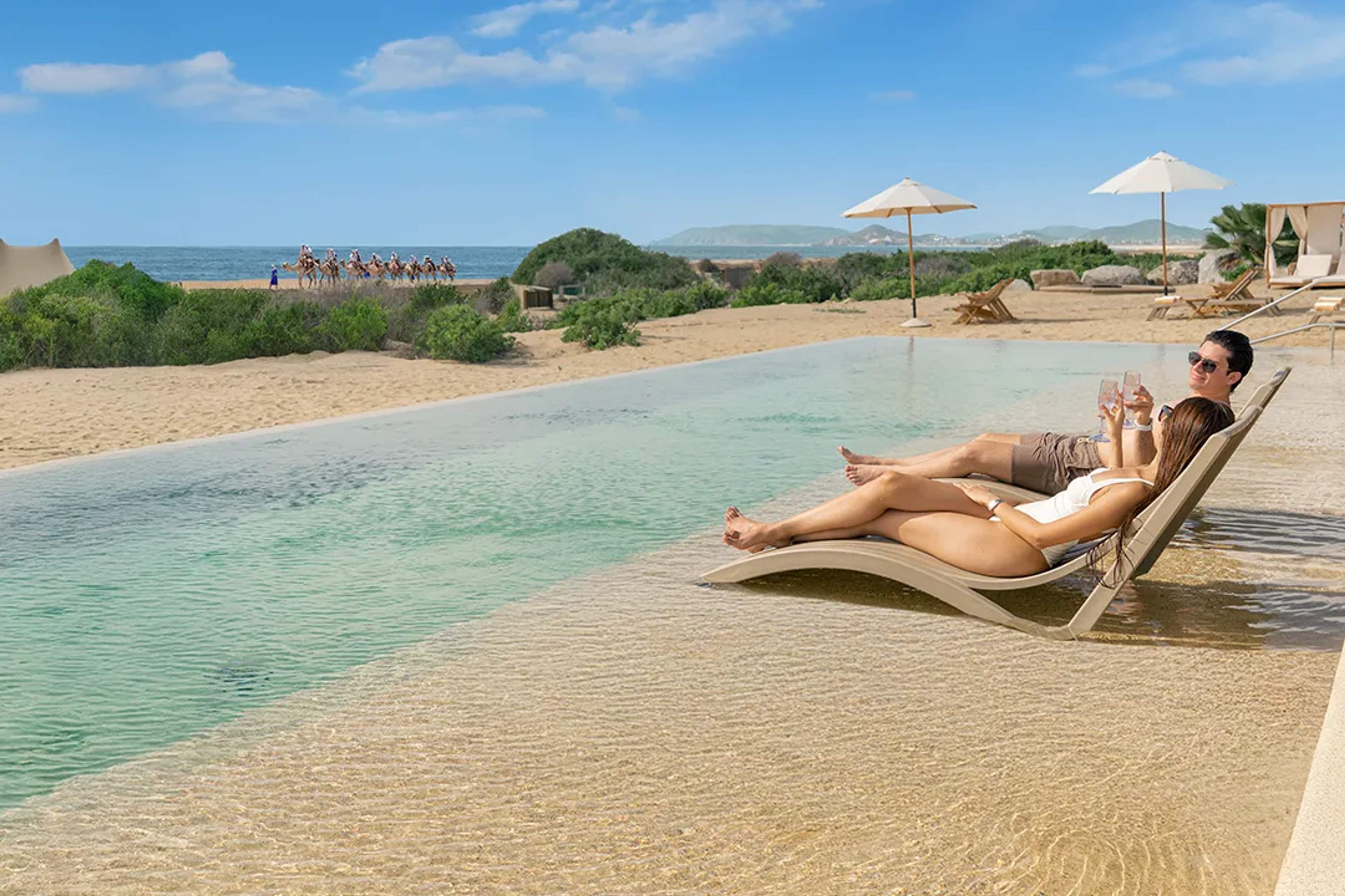 Couple relaxing in beachfront infinity pool in Cabo, sipping drinks with camels passing along the shore.