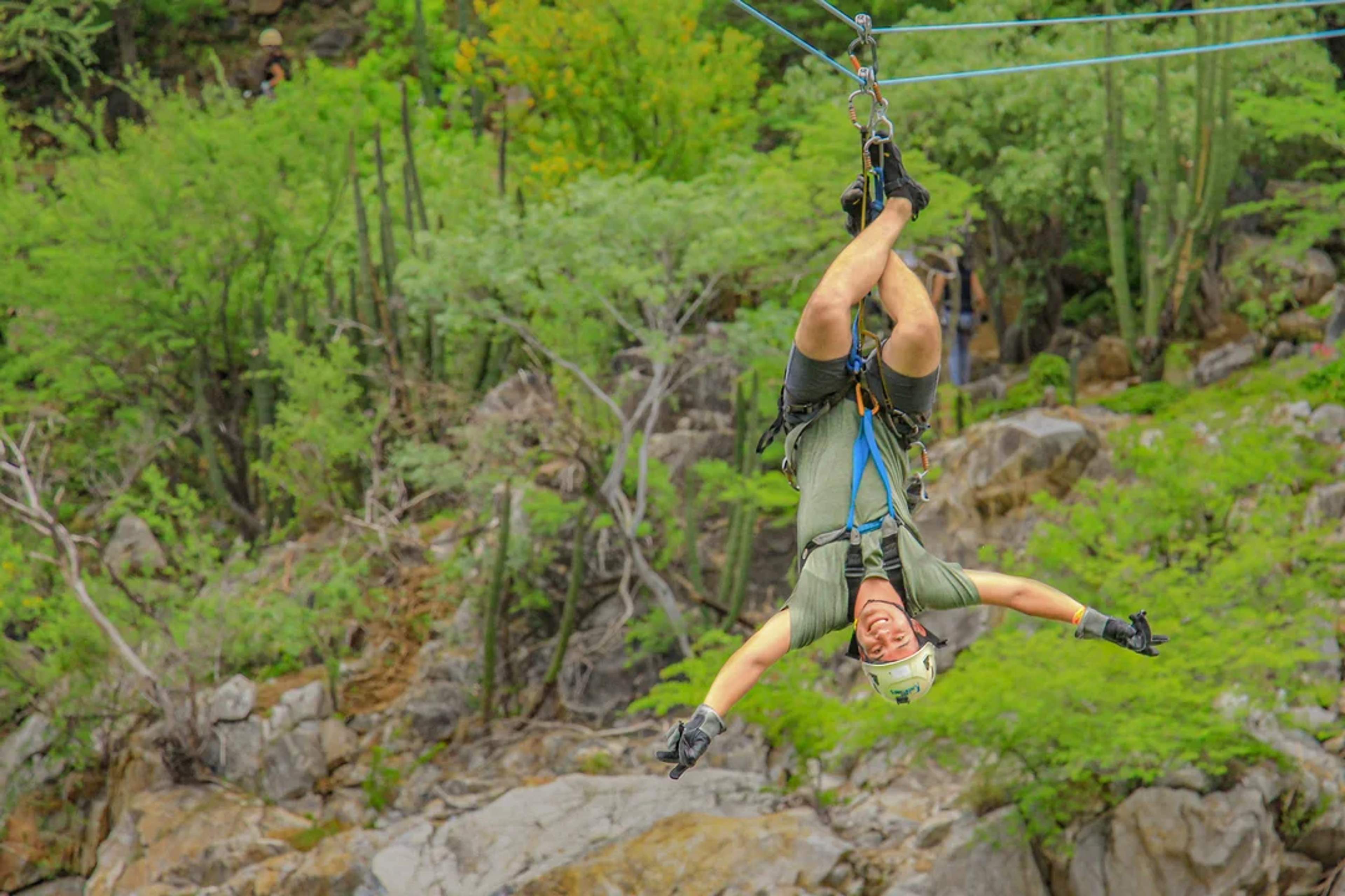 Man rides a zipline upside down with a big smile, surrounded by desert greenery and rocks.