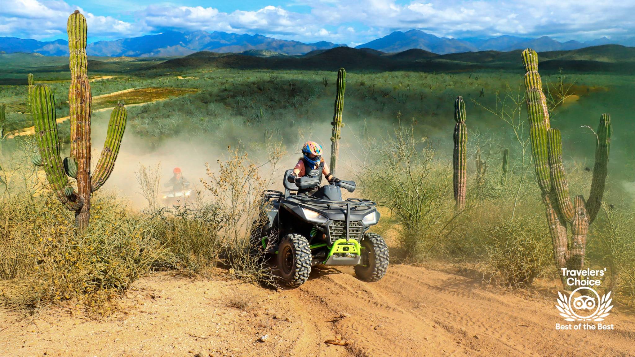 ATV rider on a dusty desert trail surrounded by cacti, mountains in the background, with a Travelers' Choice badge displayed.