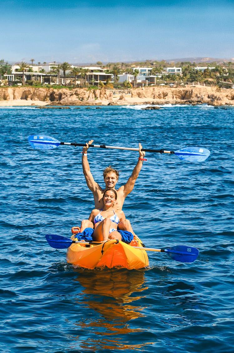 Una pareja en kayak en el océano, sonriendo y levantando sus remos, con una costa rocosa y casas al fondo.
