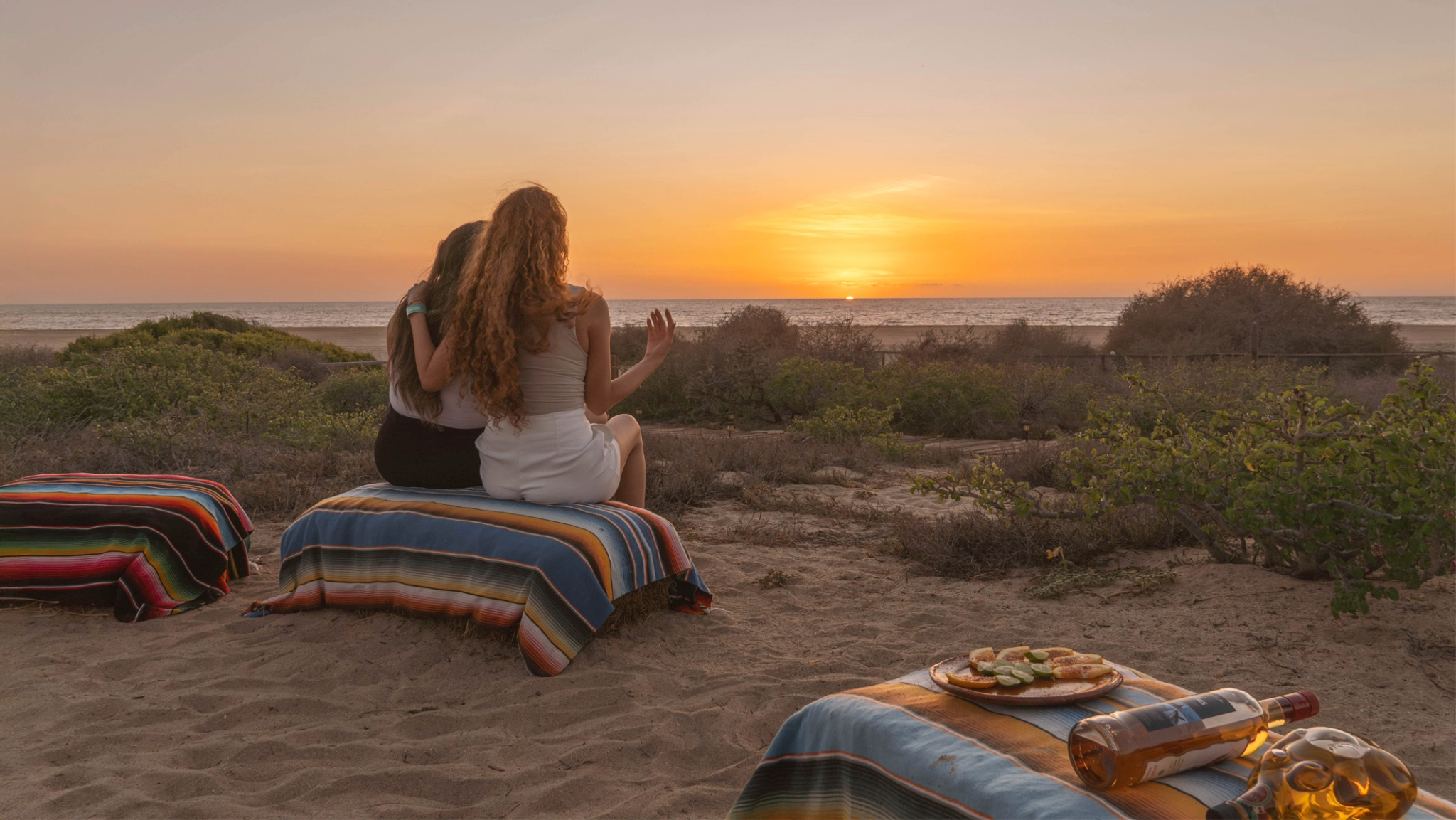 Two people sit on colorful blankets, watching the sunset over the ocean with snacks and drinks nearby on a sandy beach.