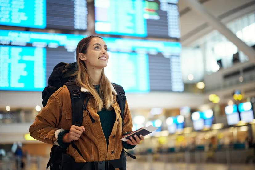 Smiling traveler with a backpack holds passport and ticket while looking at flight boards.