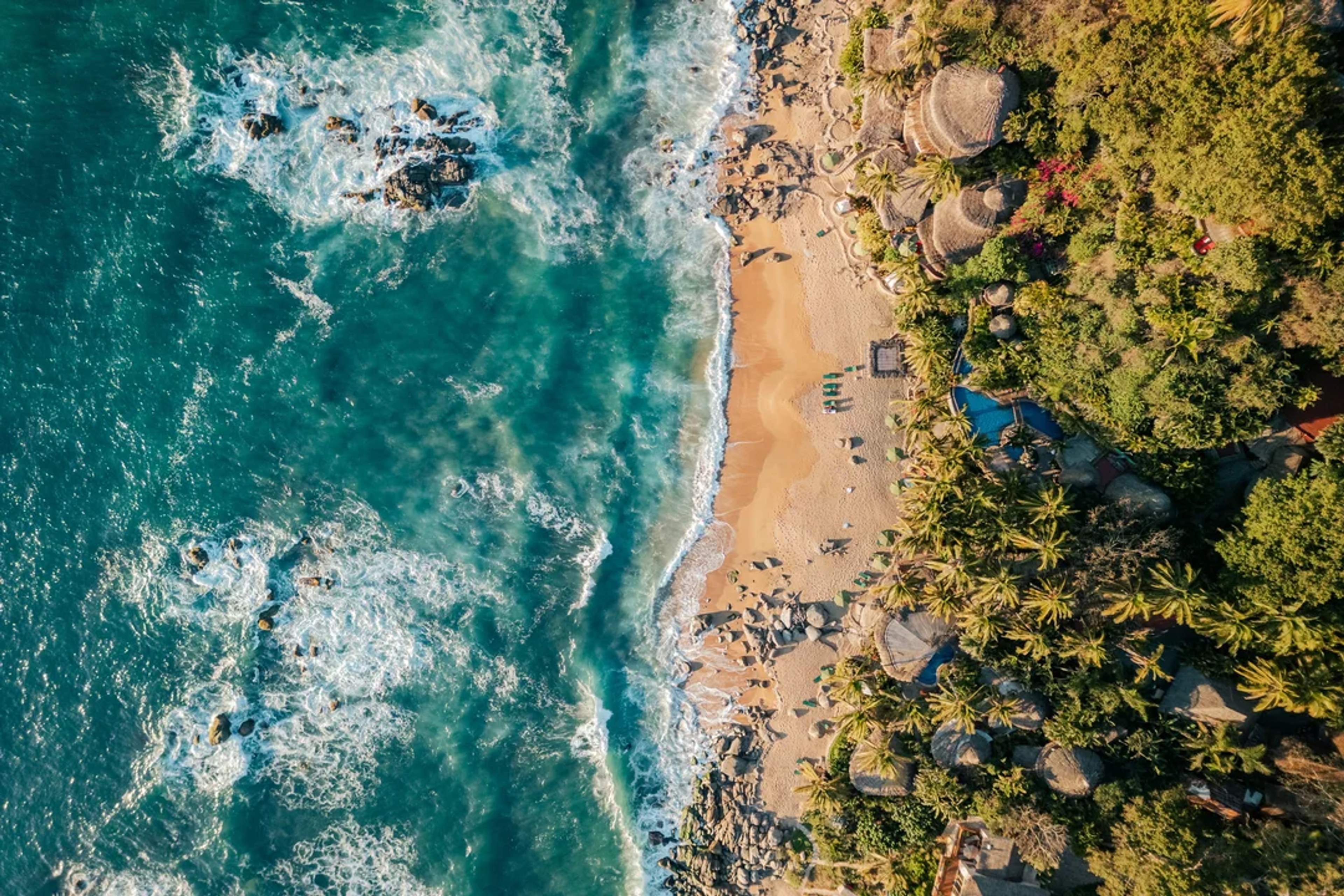 Aerial view of a tropical beach with waves, palm trees, and rustic huts nestled in lush greenery.