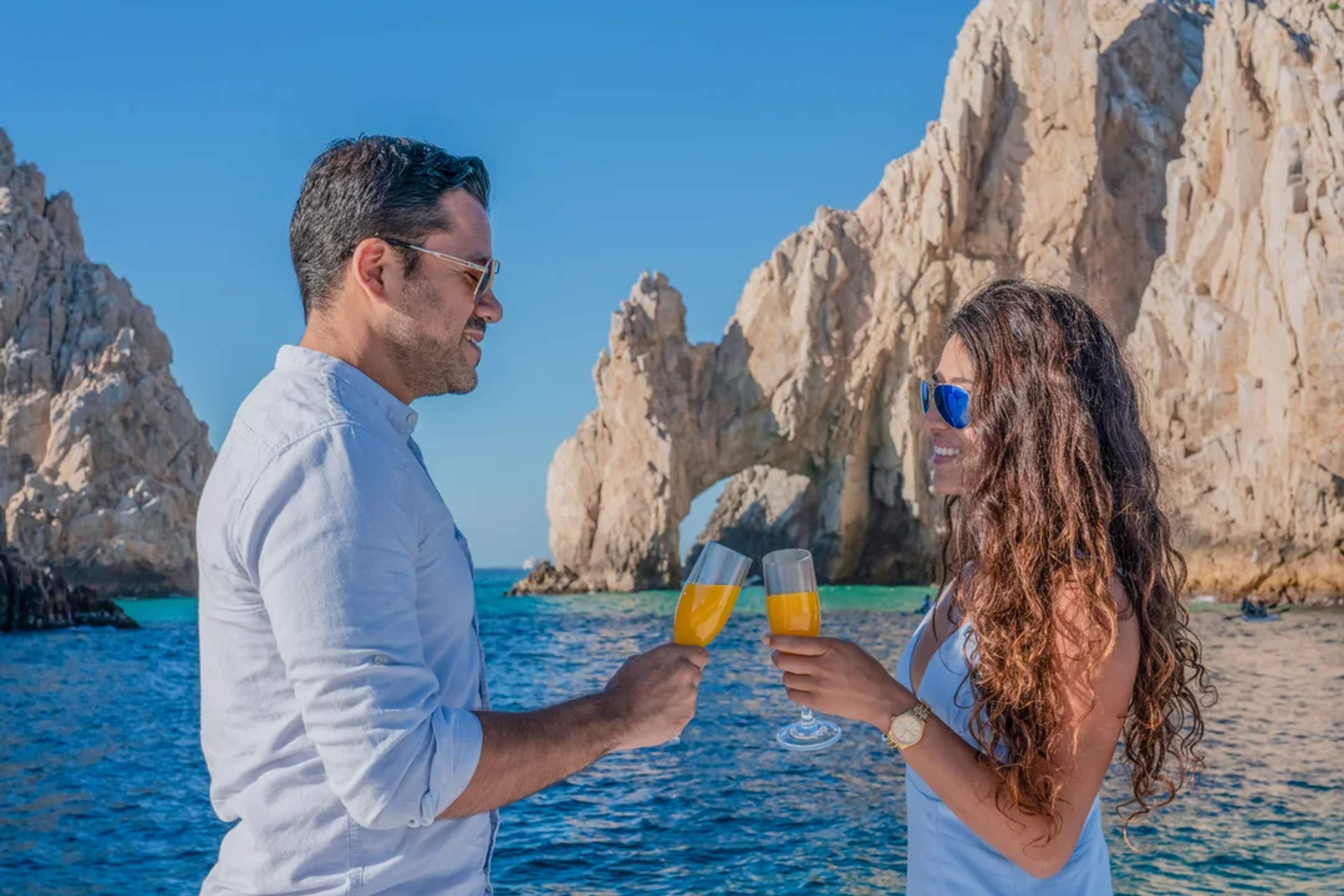 Pareja brinda con mimosas junto al Arco de Cabo, celebrando el amor bajo el sol de Baja.