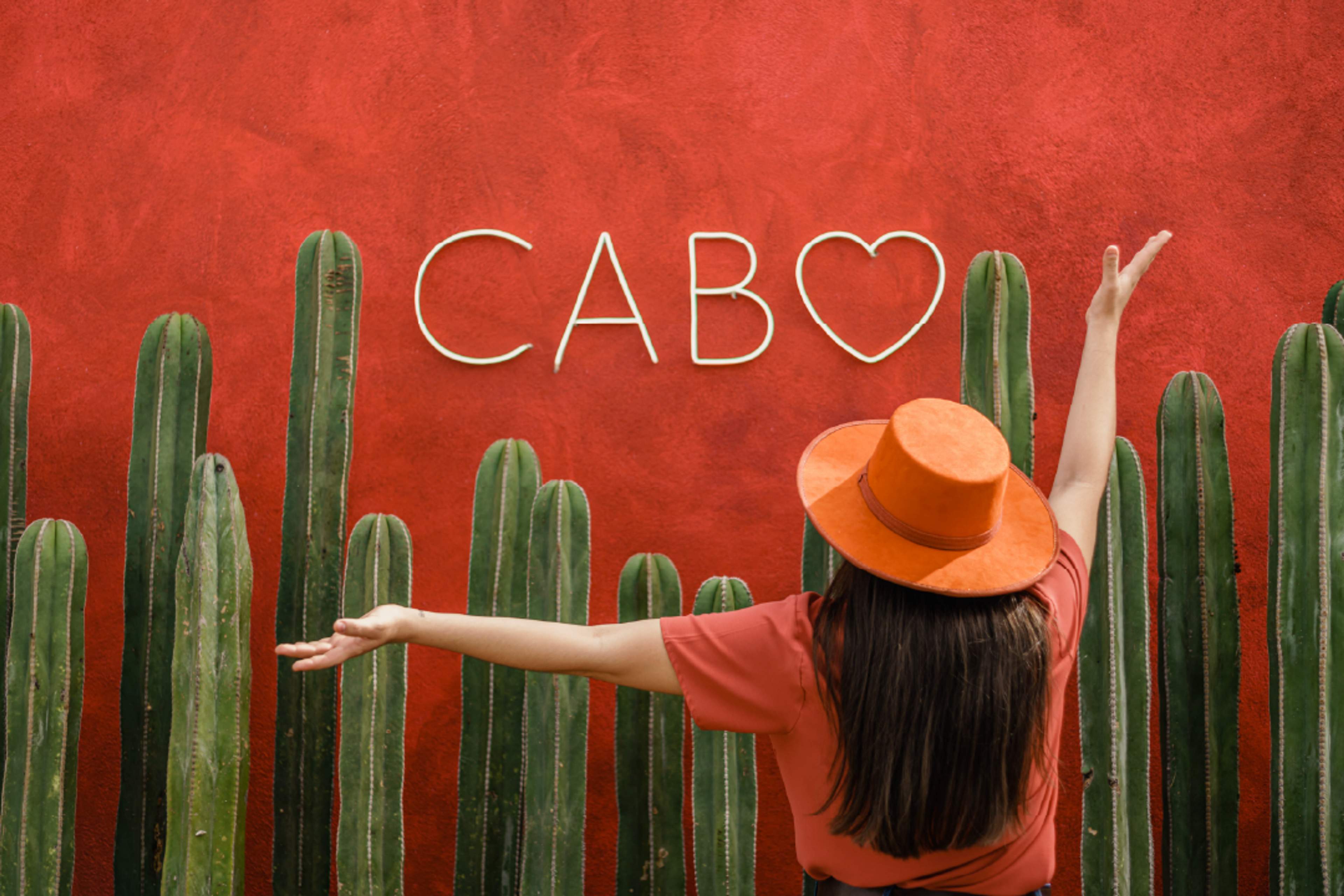 Woman in orange hat posing at the iconic Cabo sign with cacti wall, Instagram spot in Los Cabos