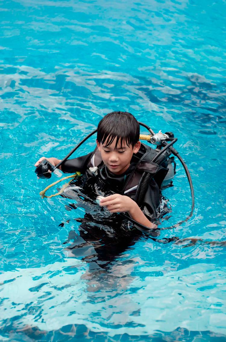 Young diver in a pool inspecting scuba gear while floating in clear blue water.