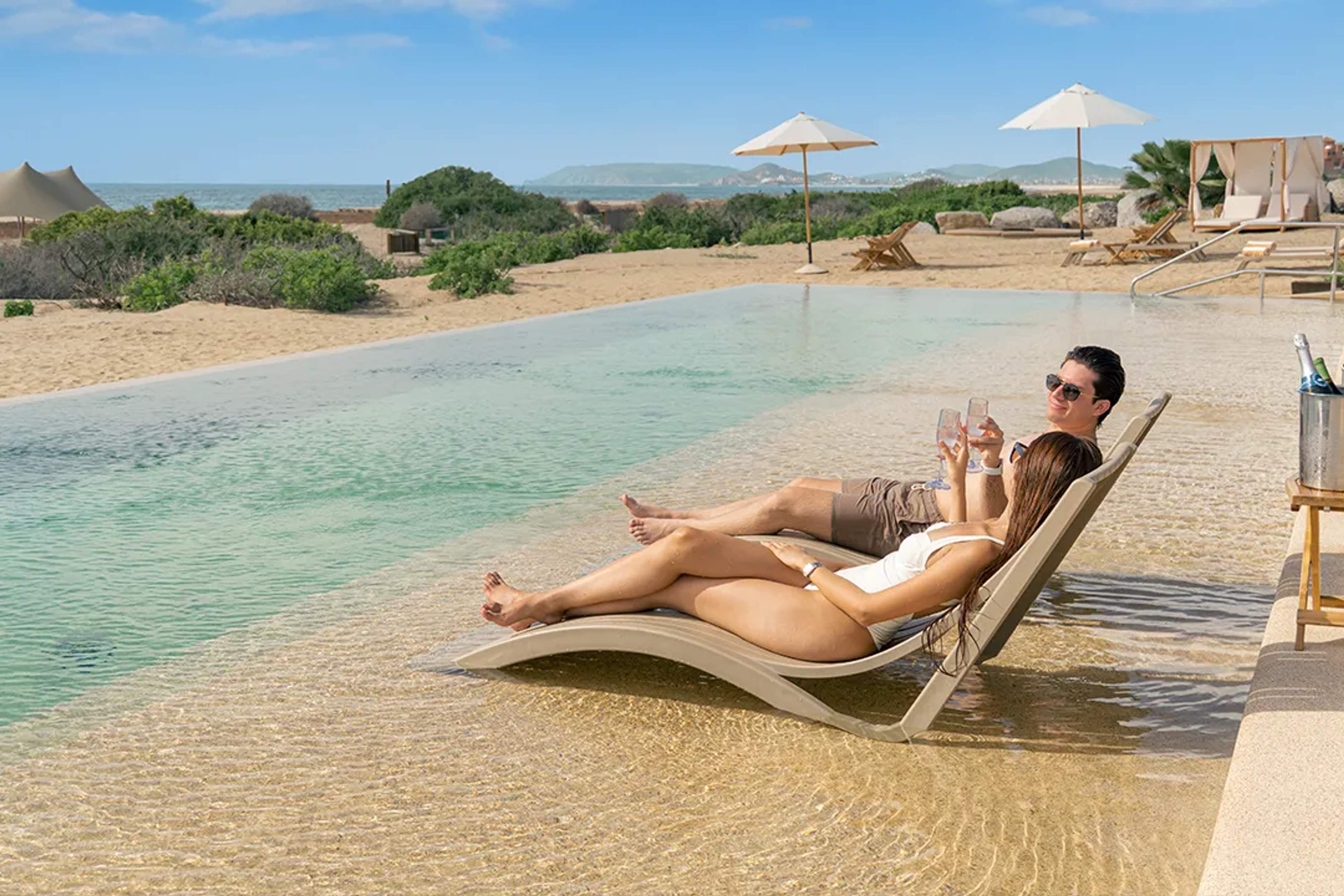 Couple relaxing on in-pool loungers with drinks at a luxury beach club in Cabo, enjoying a sunny getaway