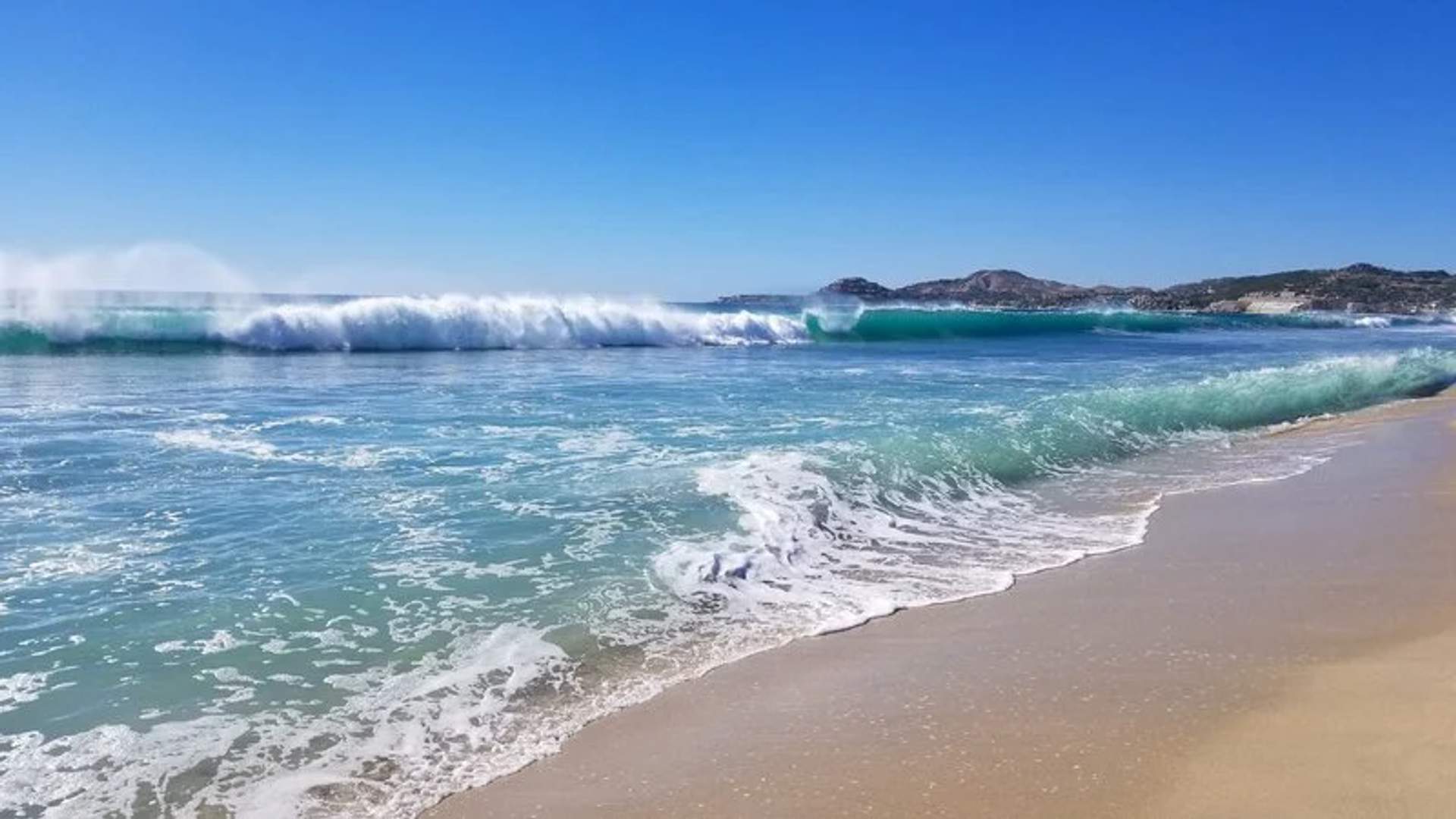 La imagen muestra una hermosa playa con olas azules claras rompiendo suavemente sobre la orilla de arena, típica de las costas escénicas en Los Cabos.
