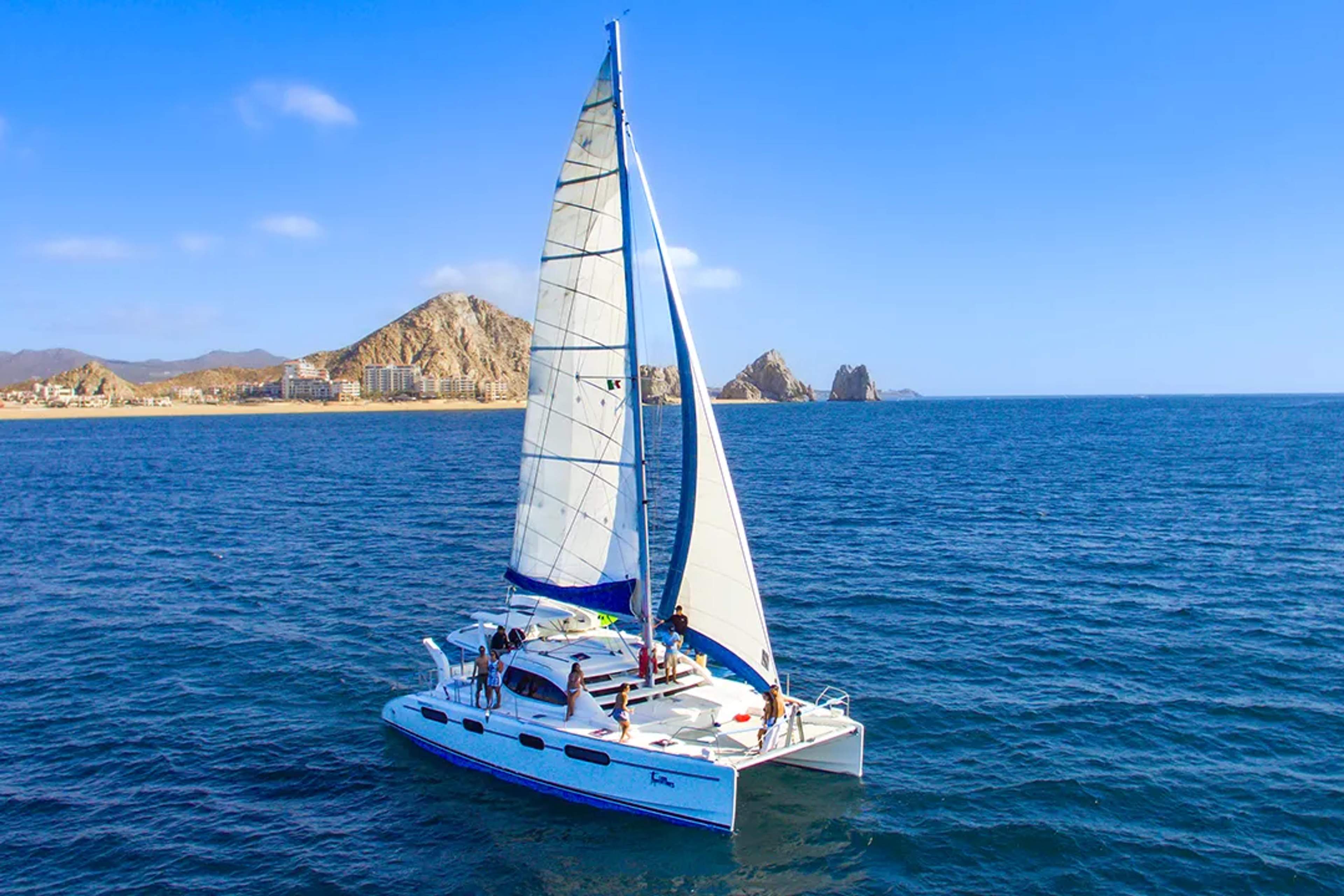 Catamaran sailing in Cabo San Lucas with guests onboard, blue ocean views and iconic rock formations ahead