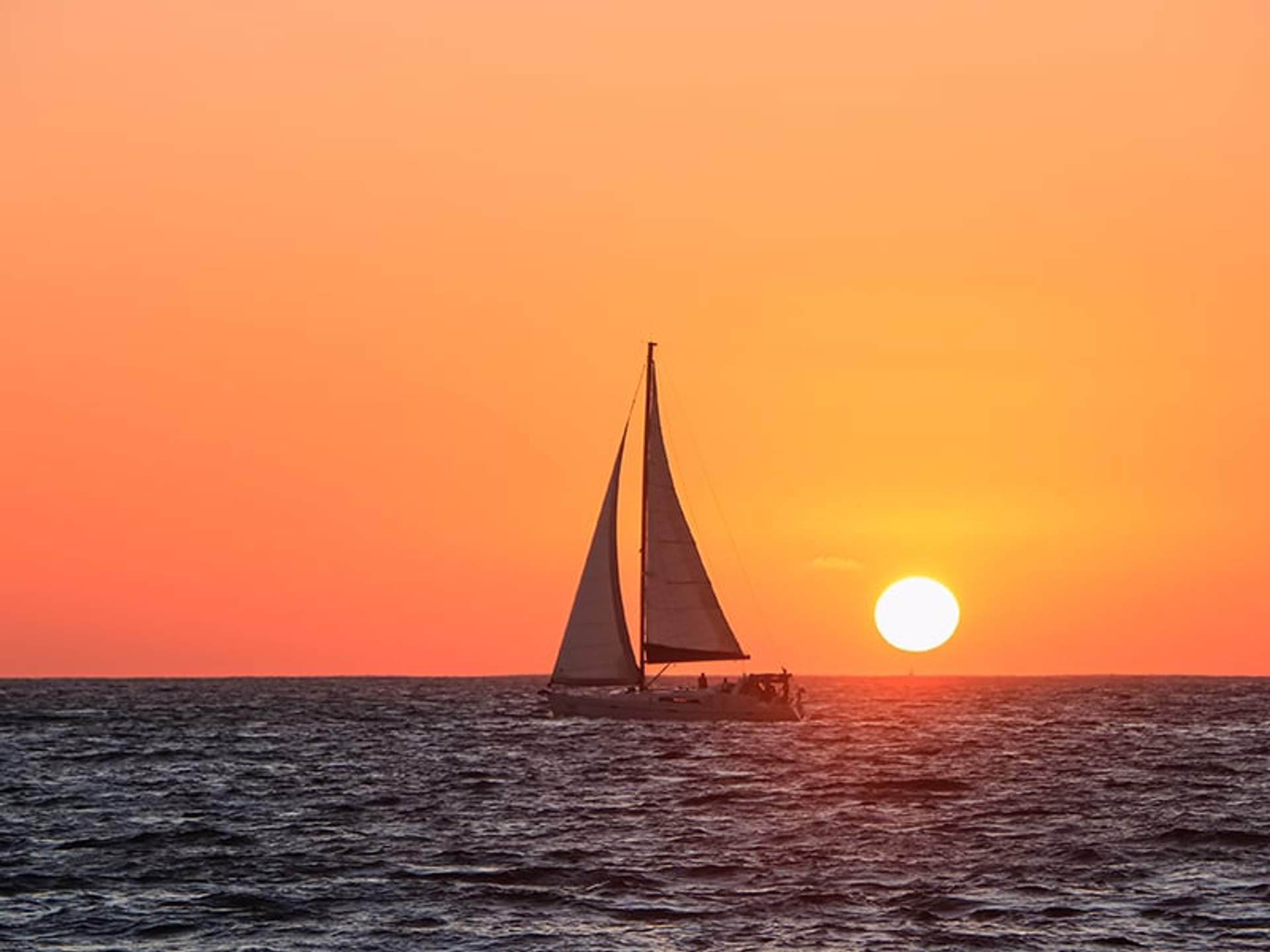 A sailboat glides on the ocean with the sun setting on the horizon, casting an orange glow across the sky and water.
